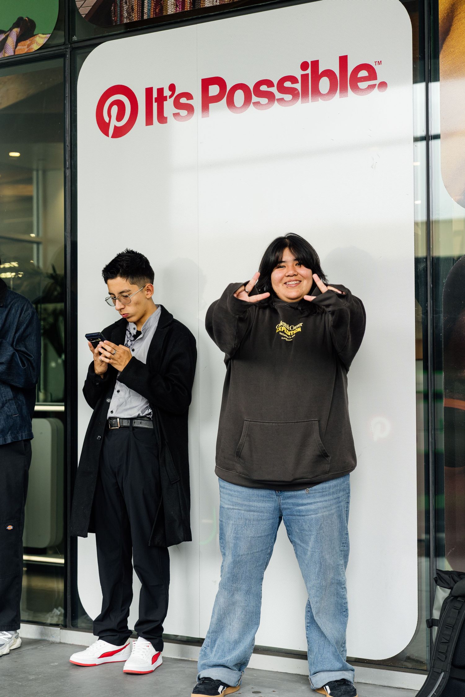 Two people posing in front of a Pinterest 'It's Possible' sign, one on phone, one making a fun gesture.