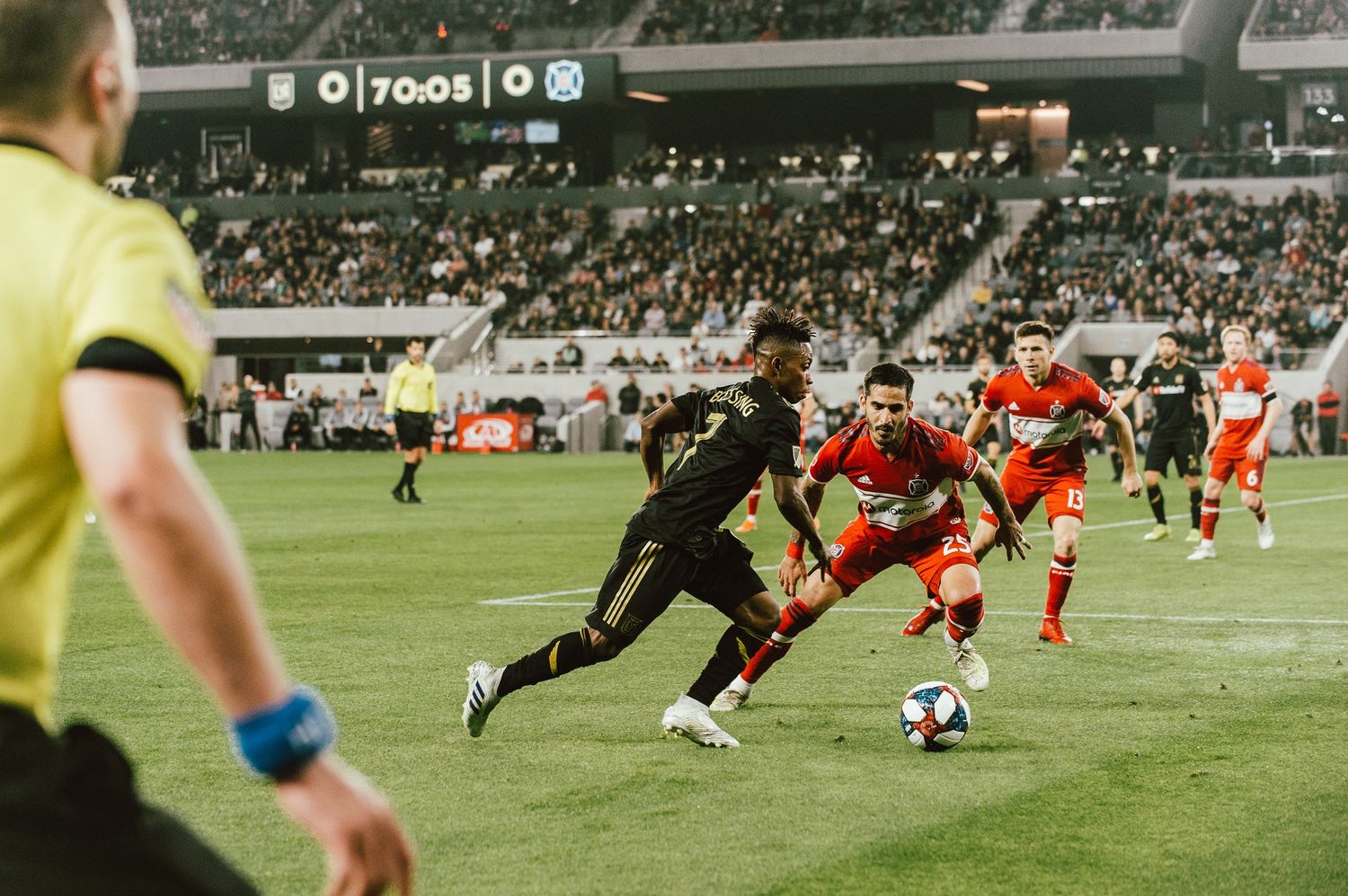 Soccer player in black uniform dribbles ball past defenders during night game at stadium.