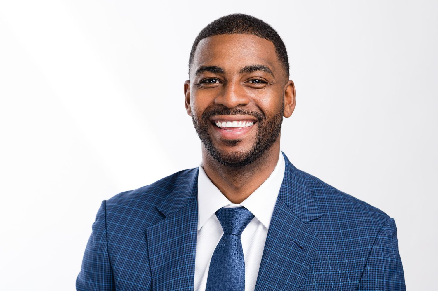 Professional headshot of someone wearing a navy checkered suit with white dress shirt and tie against light background.