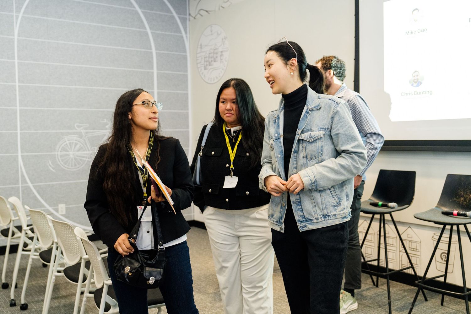 Three young women laughing and conversing at a professional networking event in a modern conference room.