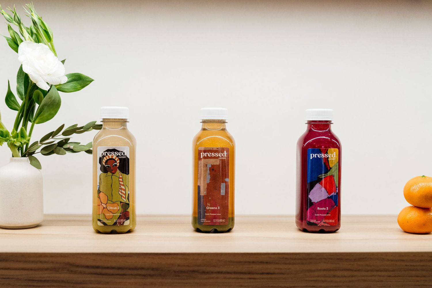 Collection of colorful juice bottles arranged on a white shelf with plants and citrus fruits.