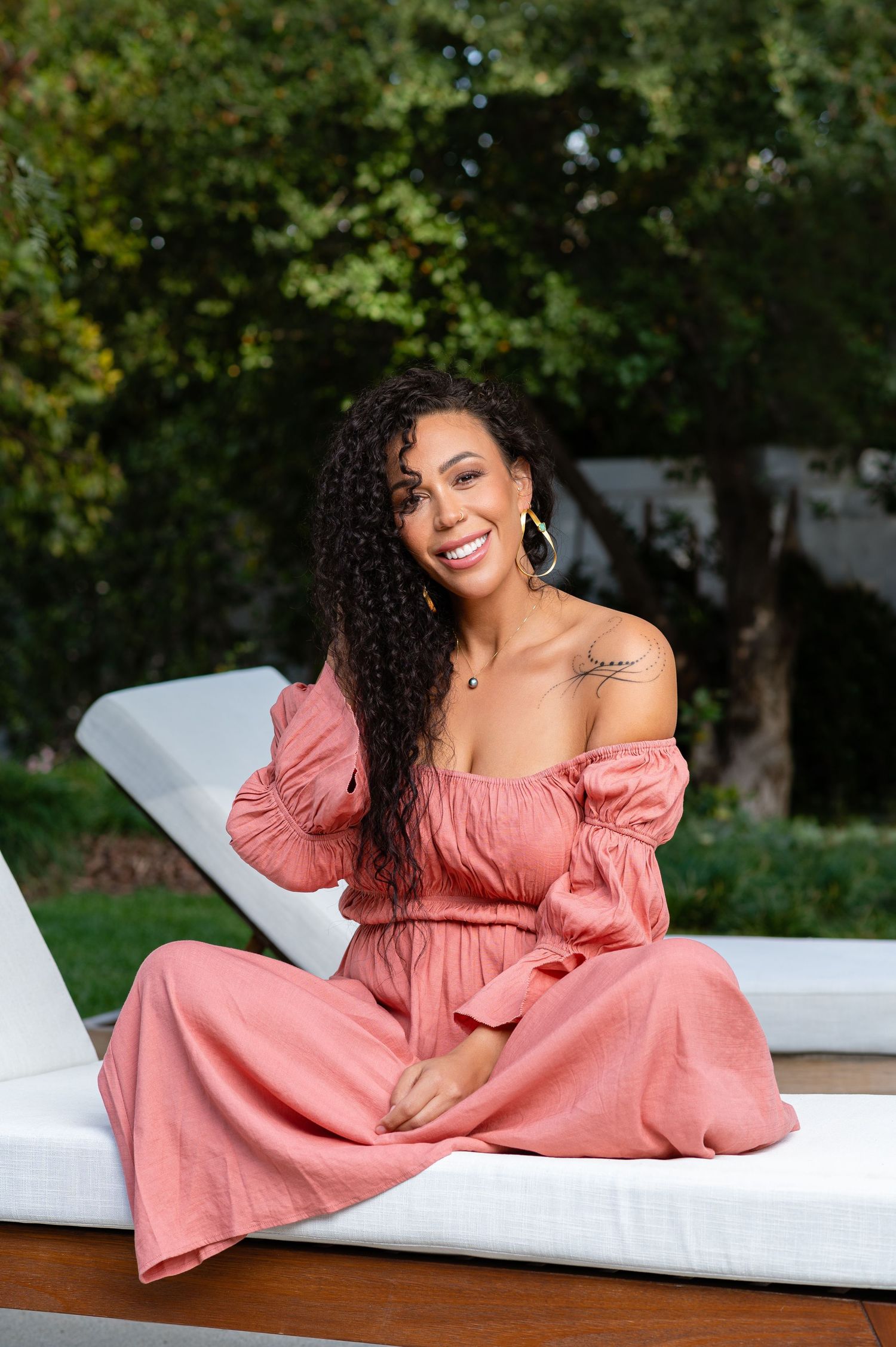 Woman in coral off-shoulder dress poses elegantly on white chaise lounge outdoors.