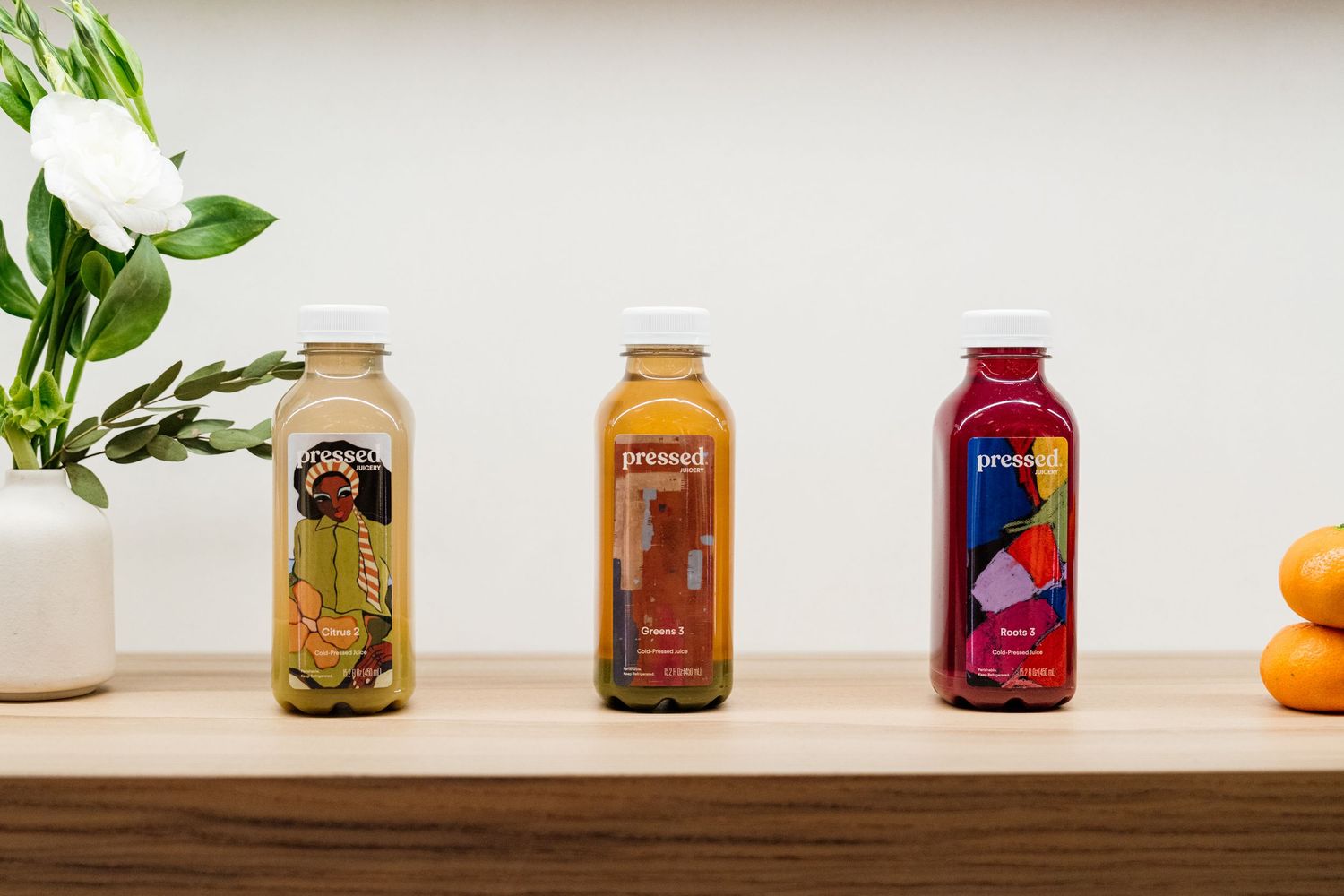 Colorful glass juice bottles arranged in a row on a white shelf with green plants in the background.