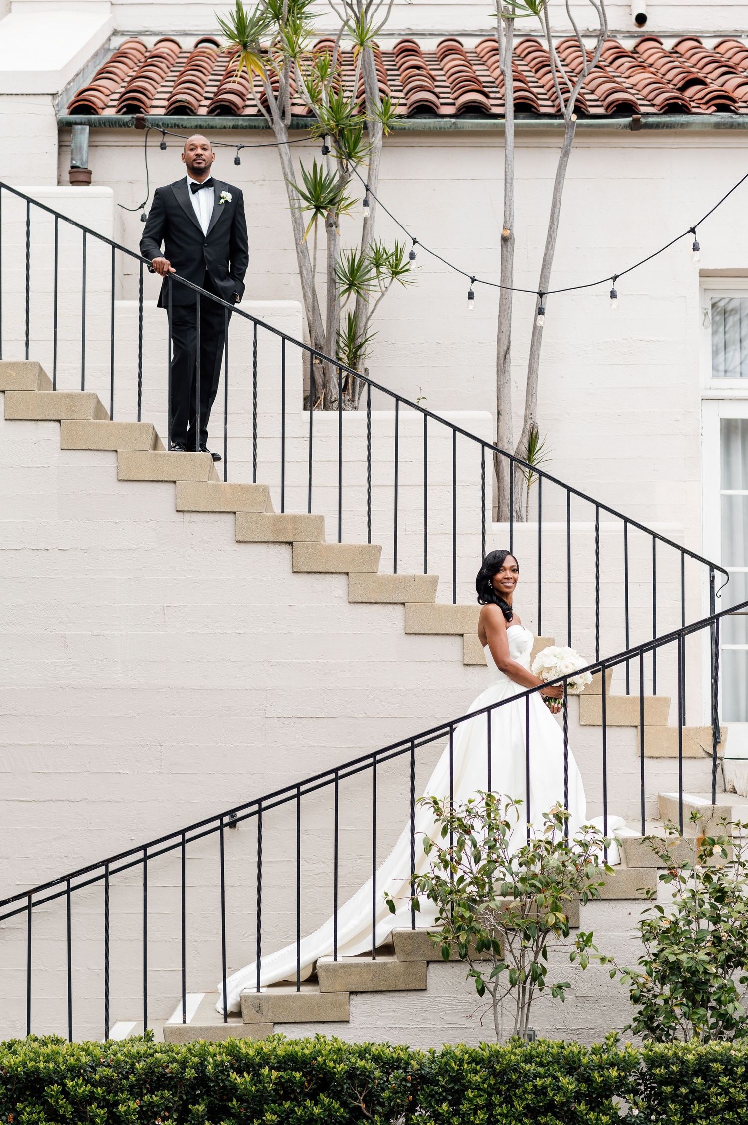 Wedding couple poses playfully on white staircase with Spanish tile roof and greenery.
