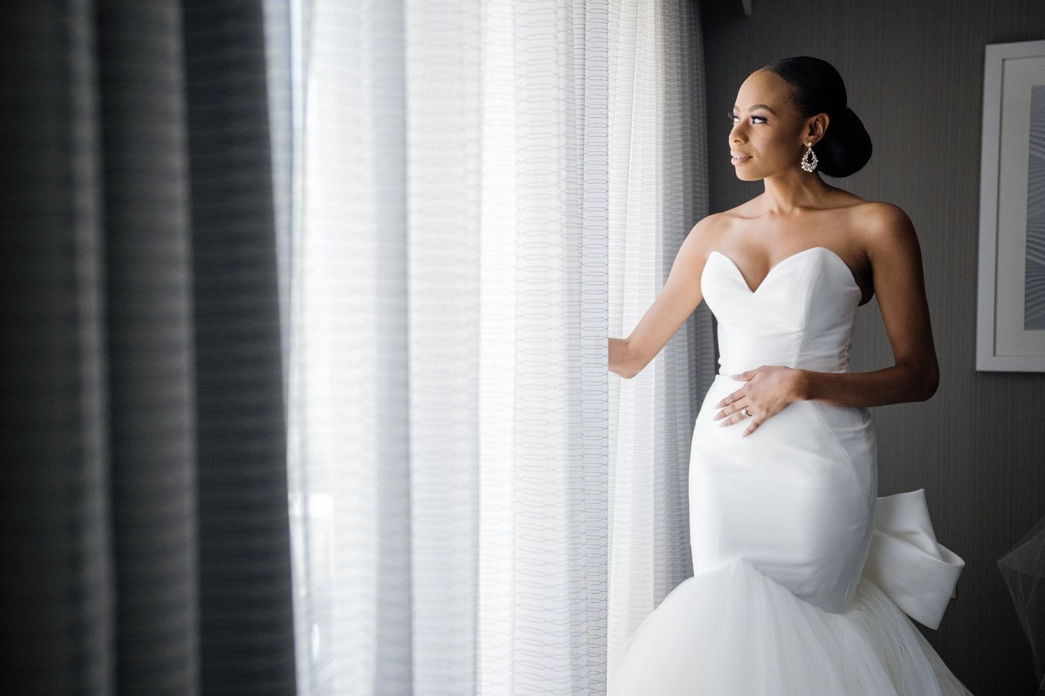 Elegant bride in white gown stands by window with dramatic lighting.