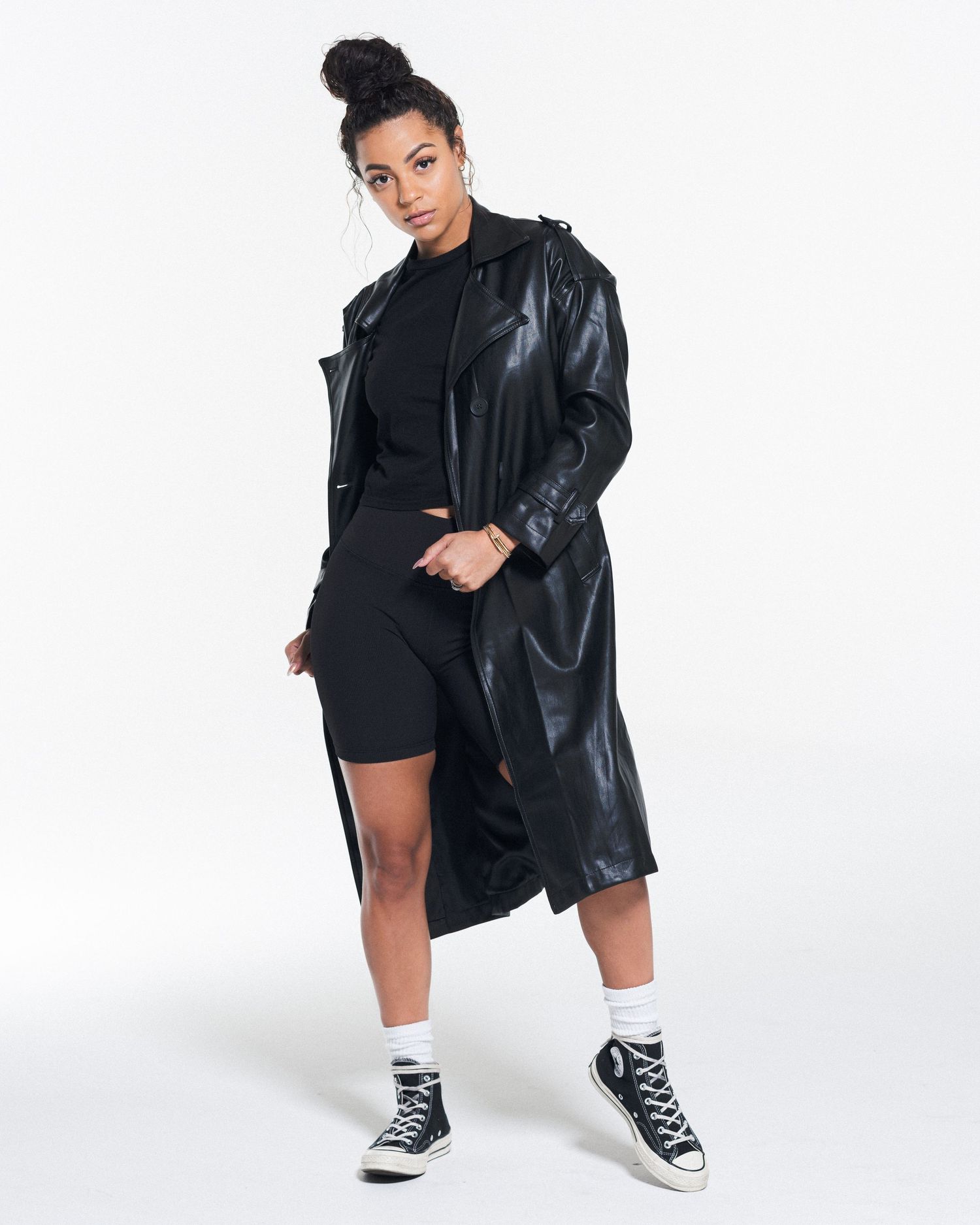 Model wearing black leather jacket and shorts with classic sneakers against white background.
