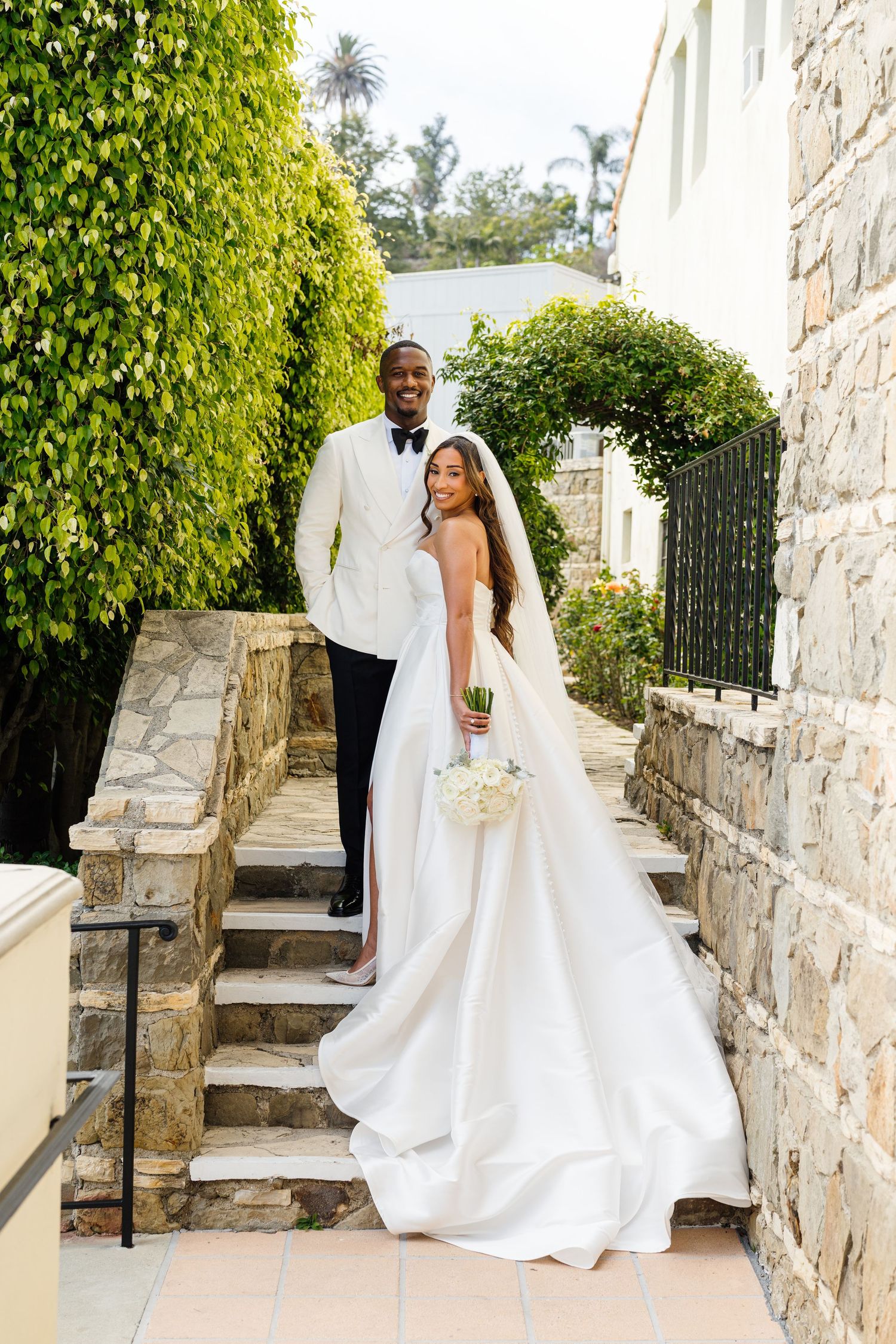 Bride in white gown holds bouquet on stone steps as groom in white tuxedo stands behind her, surrounded by lush greenery.