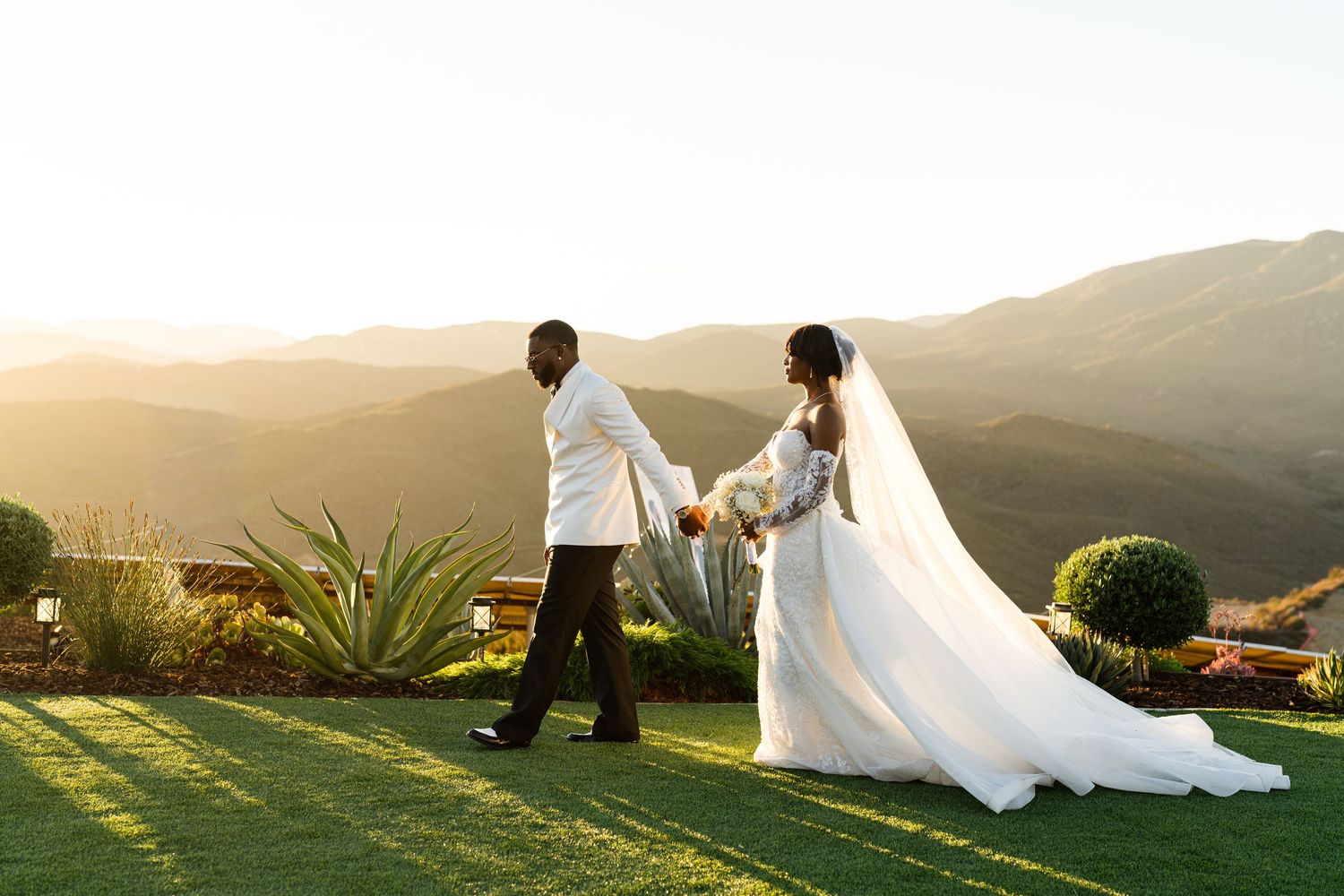 Newlywed couple holding hands at sunset with stunning mountain landscape backdrop at outdoor wedding.