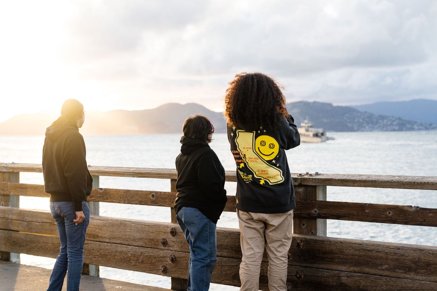 Three people stand on a wooden pier overlooking a scenic bay with mountains and sunset in the background.