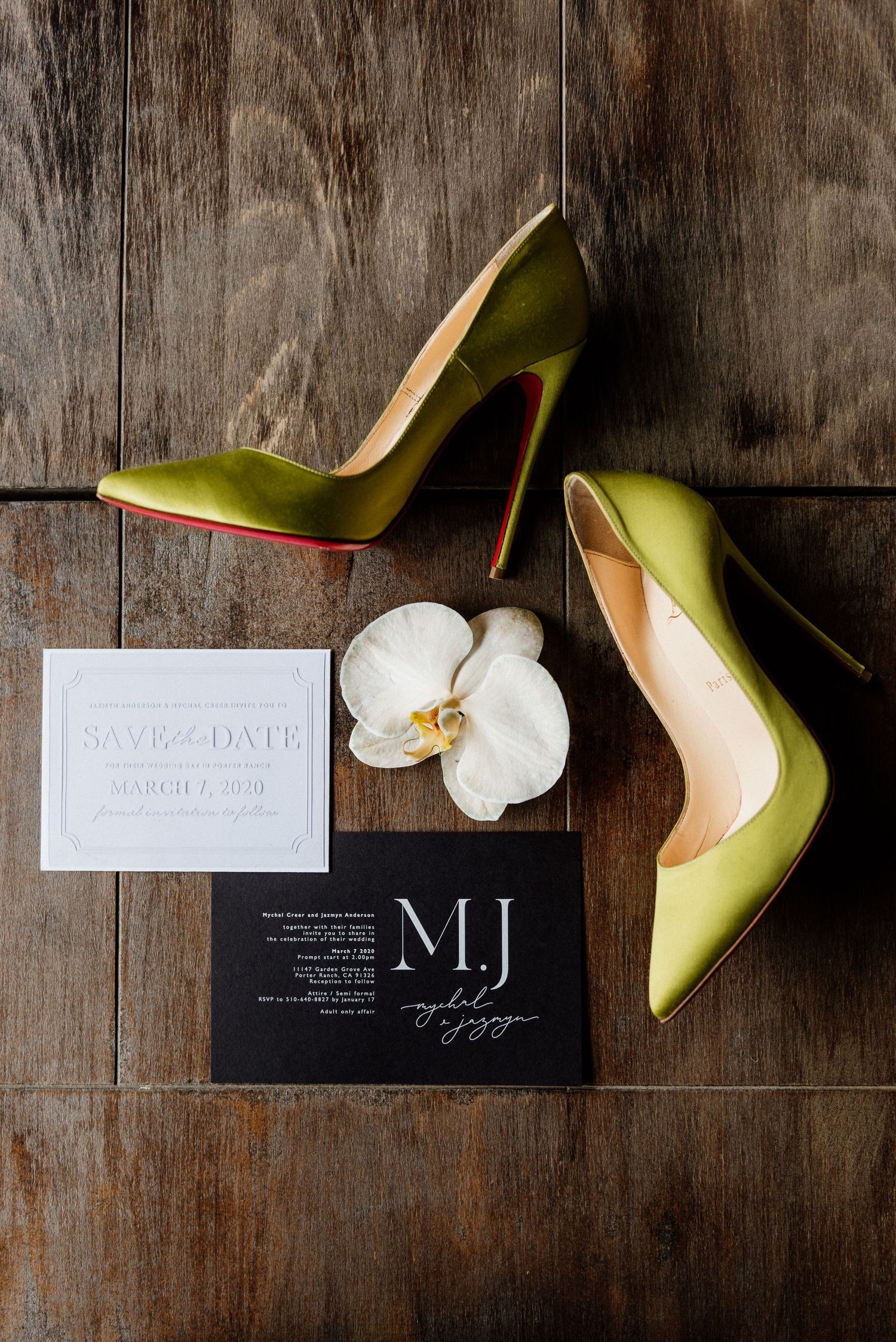 Green stiletto heels with wedding invitations and white orchid on wooden surface.