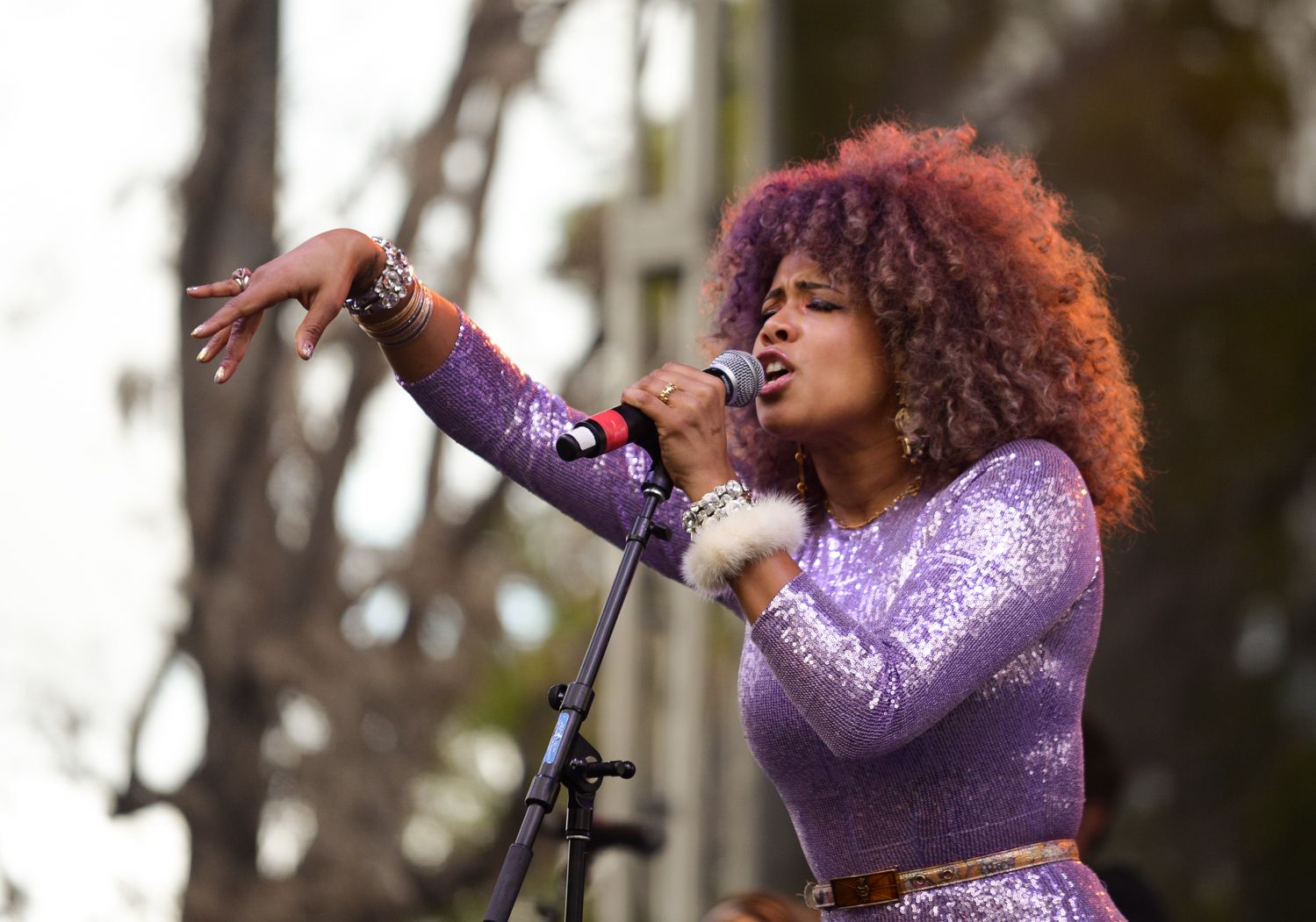 Singer in purple sequined outfit performs passionately outdoors with raised arm gesture.