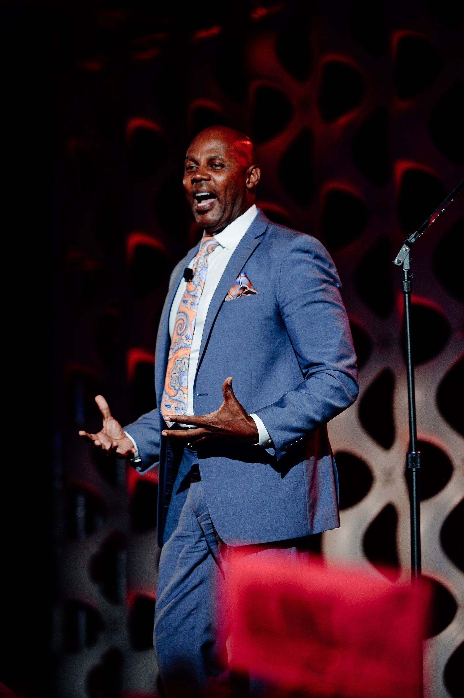 Speaker in blue suit gesturing expressively during presentation with red lighting effects.
