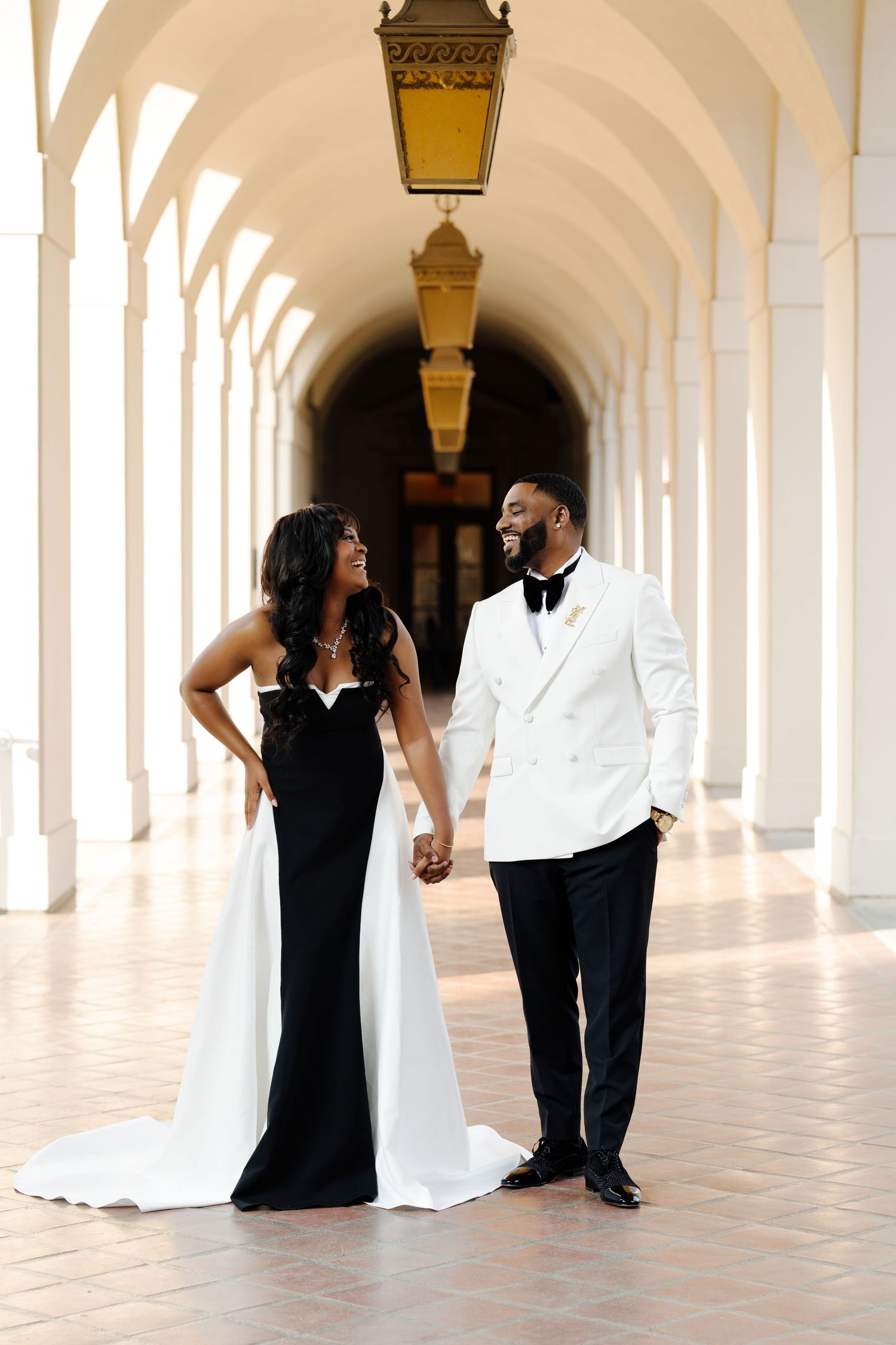 Elegant couple in formal black and white attire pose in arched colonnade hallway.