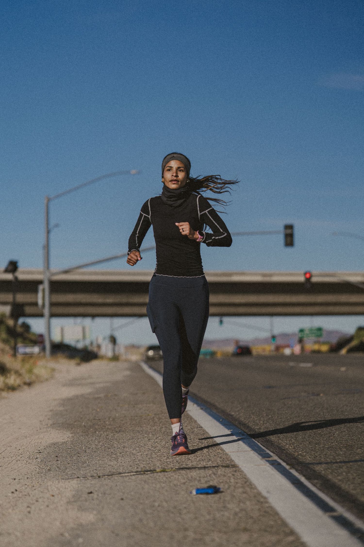 A woman in black athletic wear and hijab runs along an urban road under a clear blue sky.