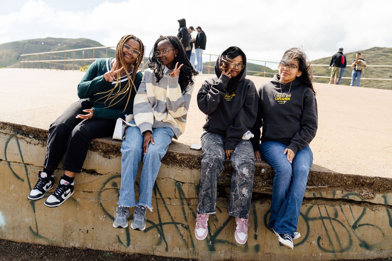 Four young women sitting on a graffiti wall outdoors, smiling and posing together on a cloudy day.