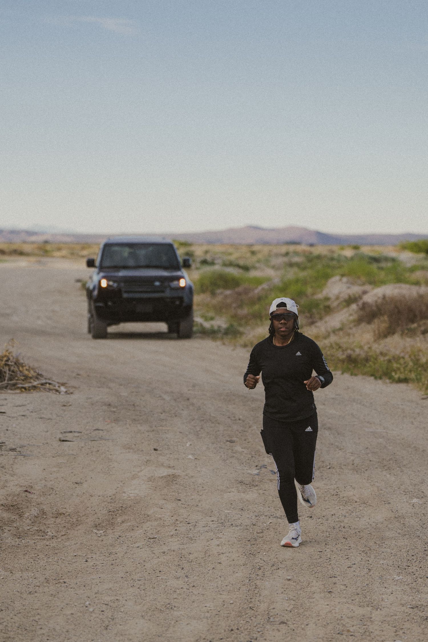Person in black Adidas outfit running on dirt road with SUV following behind in arid desert landscape.