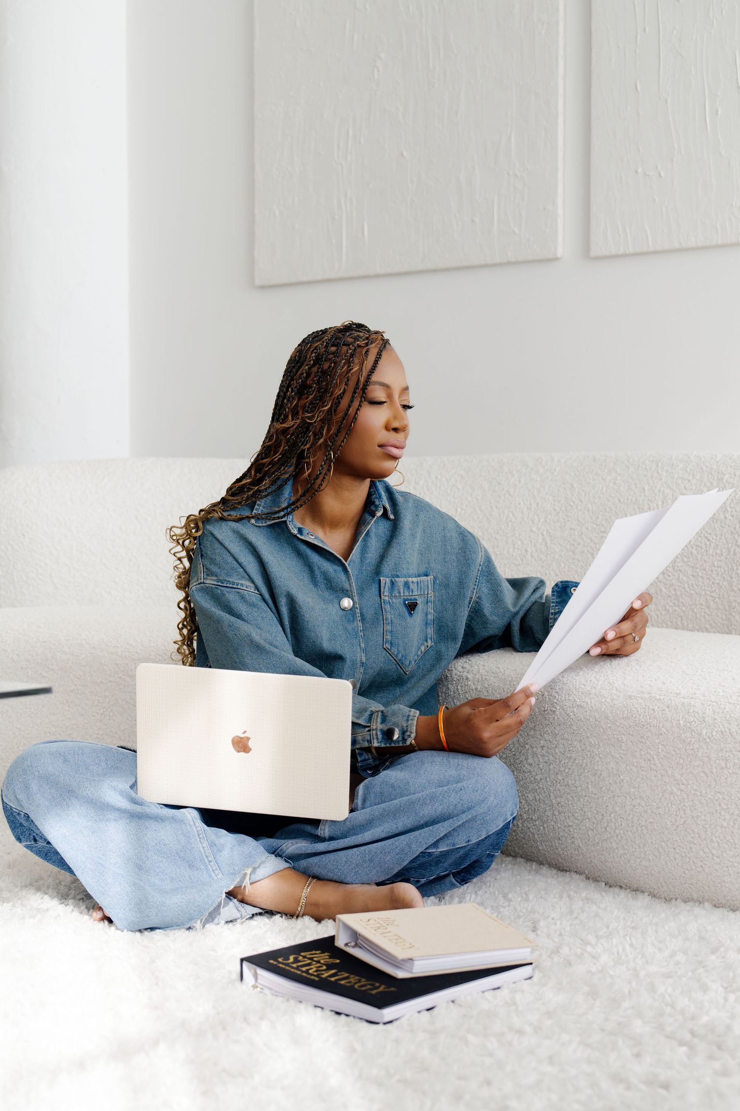 Person in denim shirt relaxes on white couch while reading documents and using laptop computer.