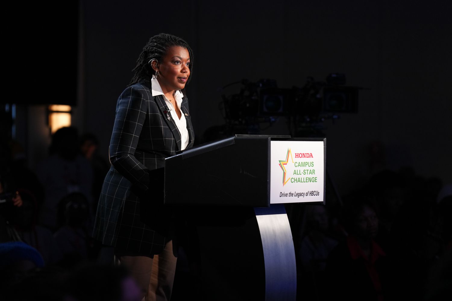 Woman in plaid blazer speaking at WNBA Campus All-Star Challenge podium during a formal event.