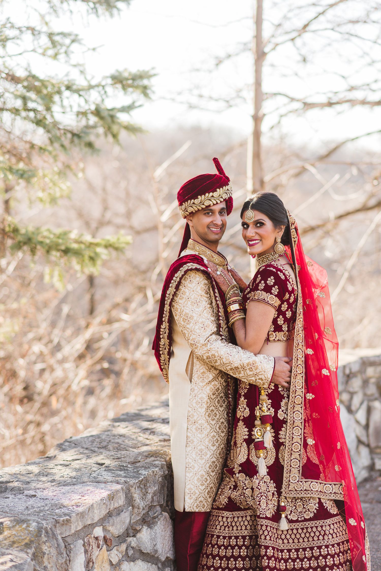 Traditional Indian wedding couple in ornate red and gold attire posing against stone wall.