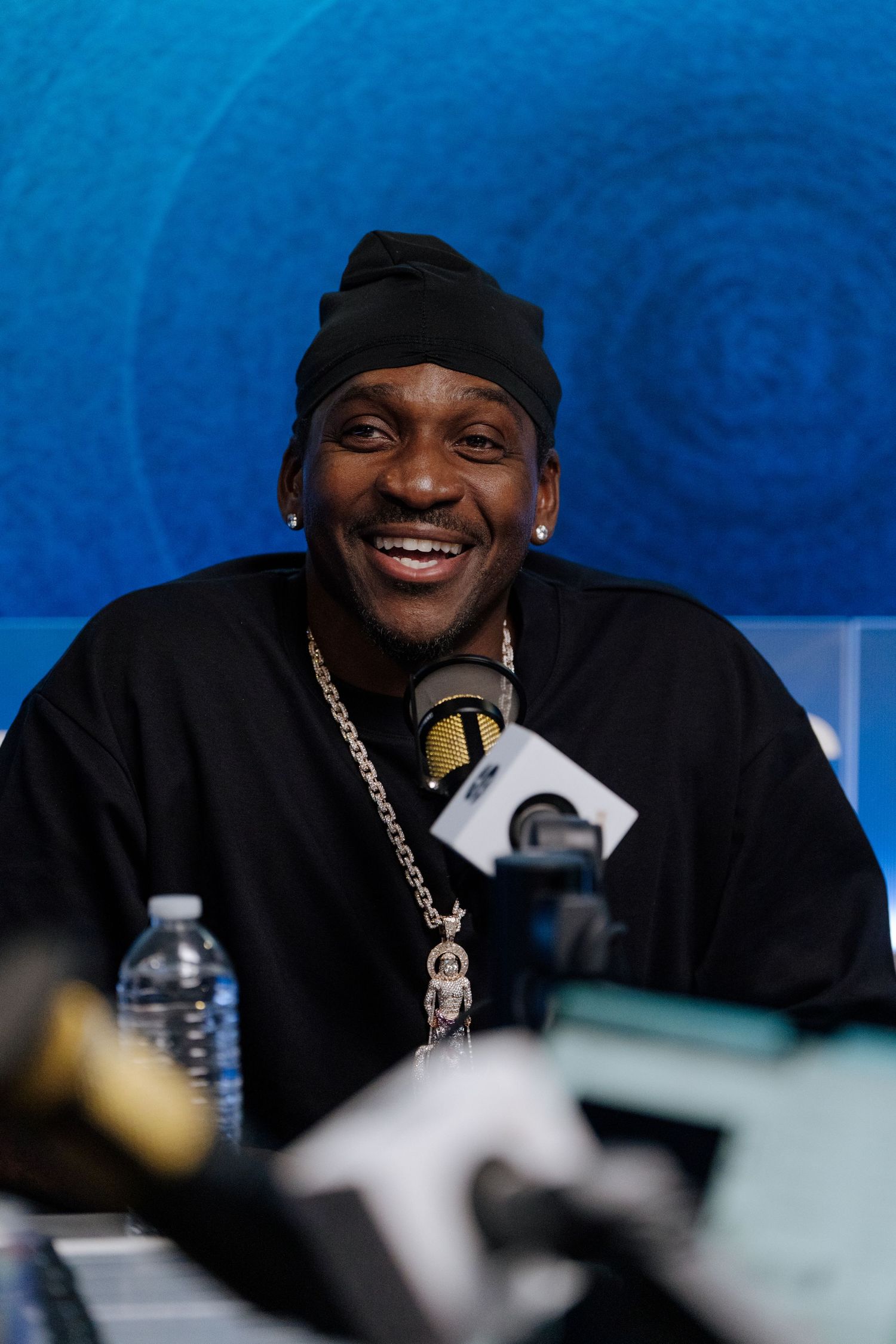 Person in black beanie and chain necklace smiling during interview against blue backdrop.