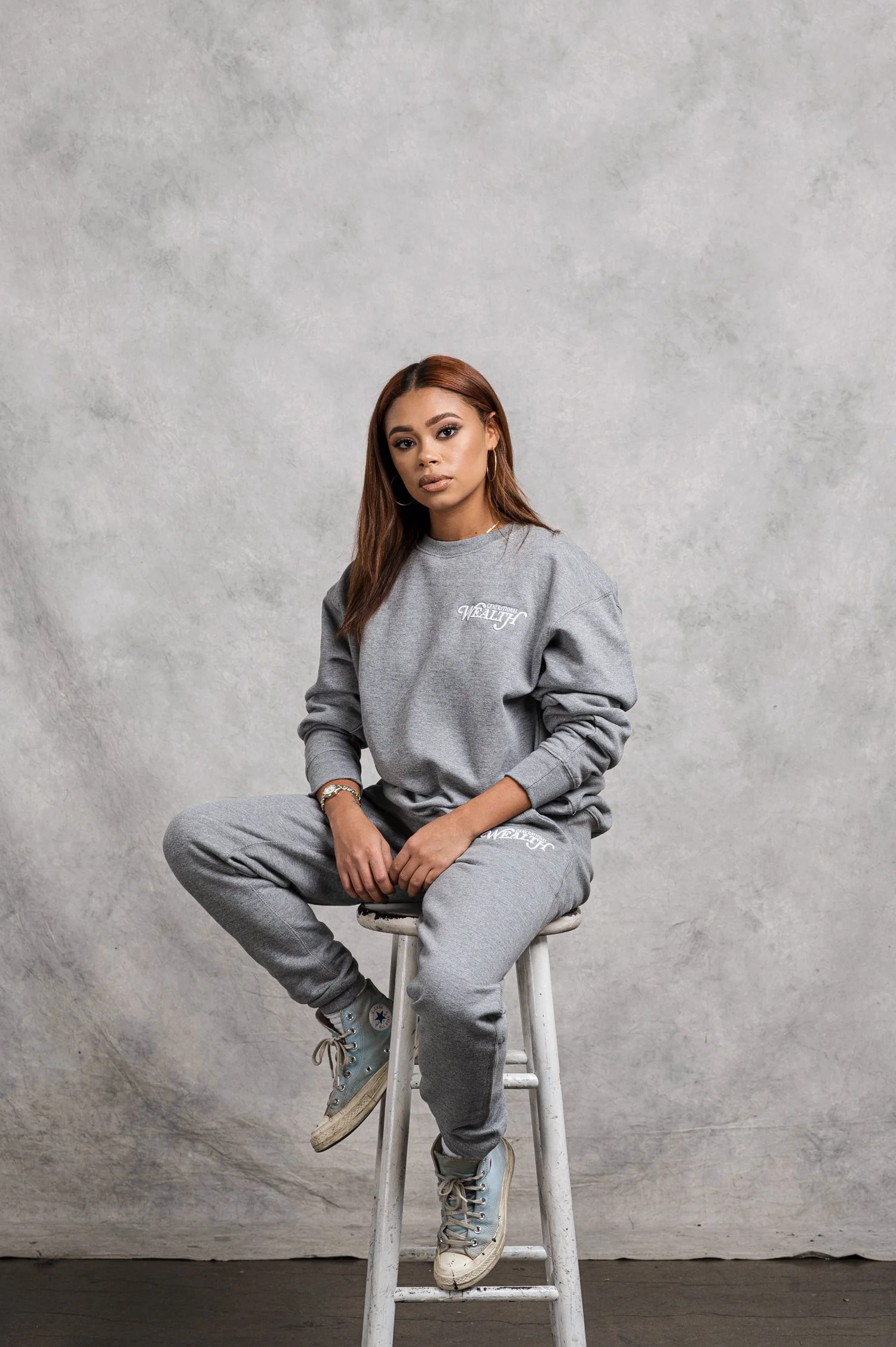 Person in grey sweatsuit lounging on stool against textured backdrop.