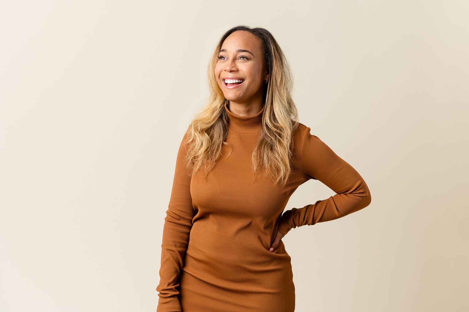 Woman in brown sweater poses confidently against cream background.