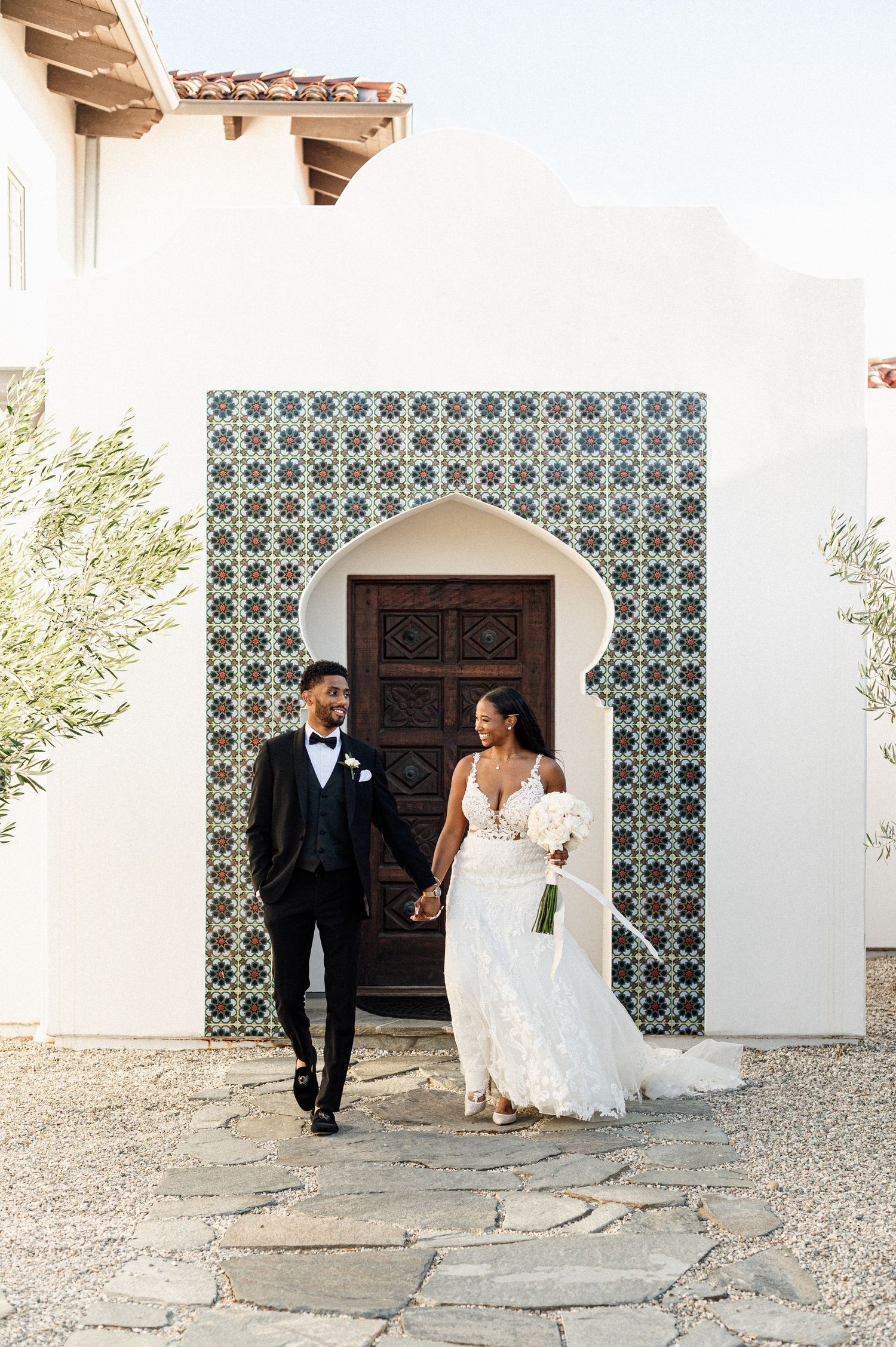 Wedding couple walks along Spanish-style building with decorative tile walls.