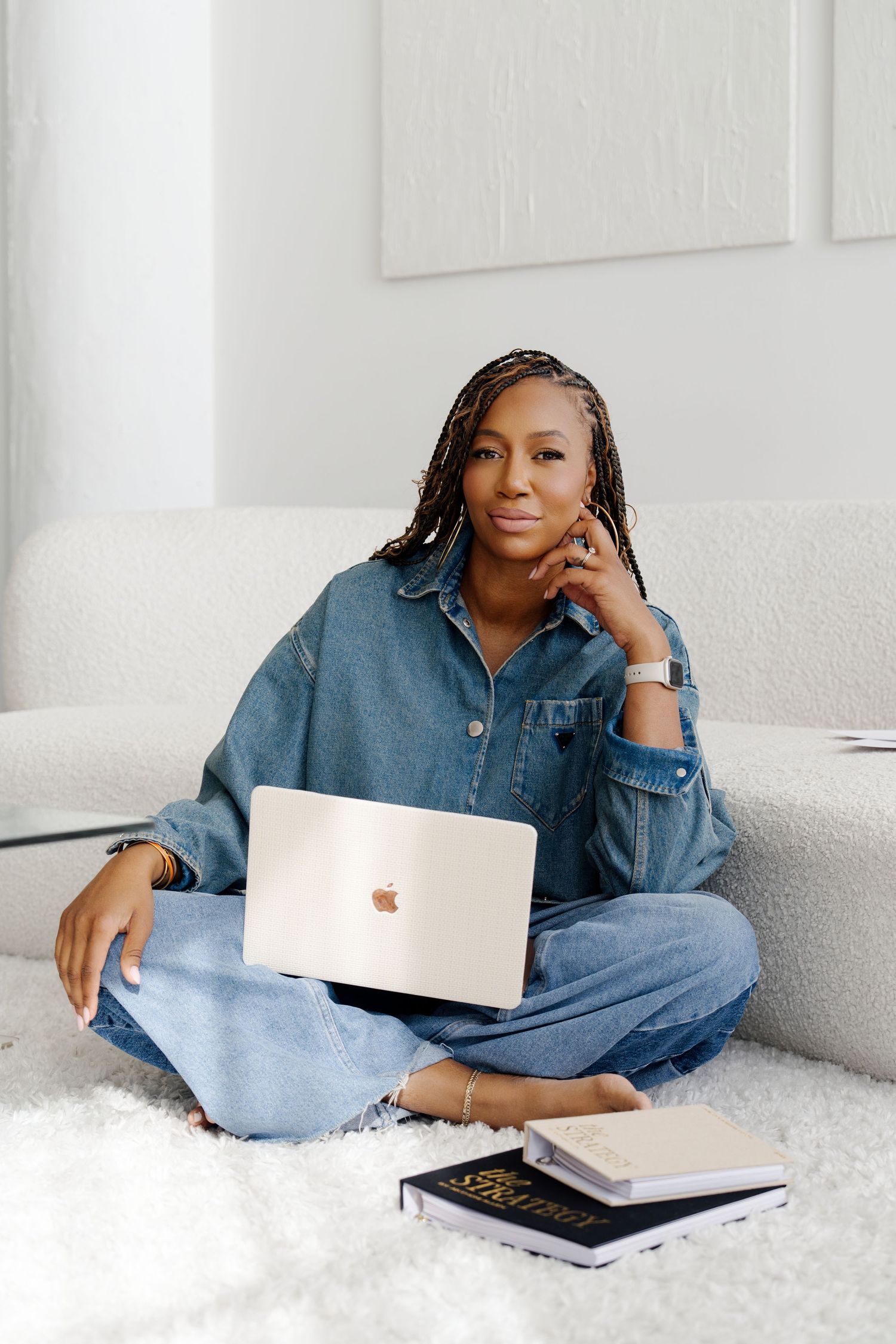Woman in denim shirt works on MacBook while sitting cross-legged on white couch with books nearby.