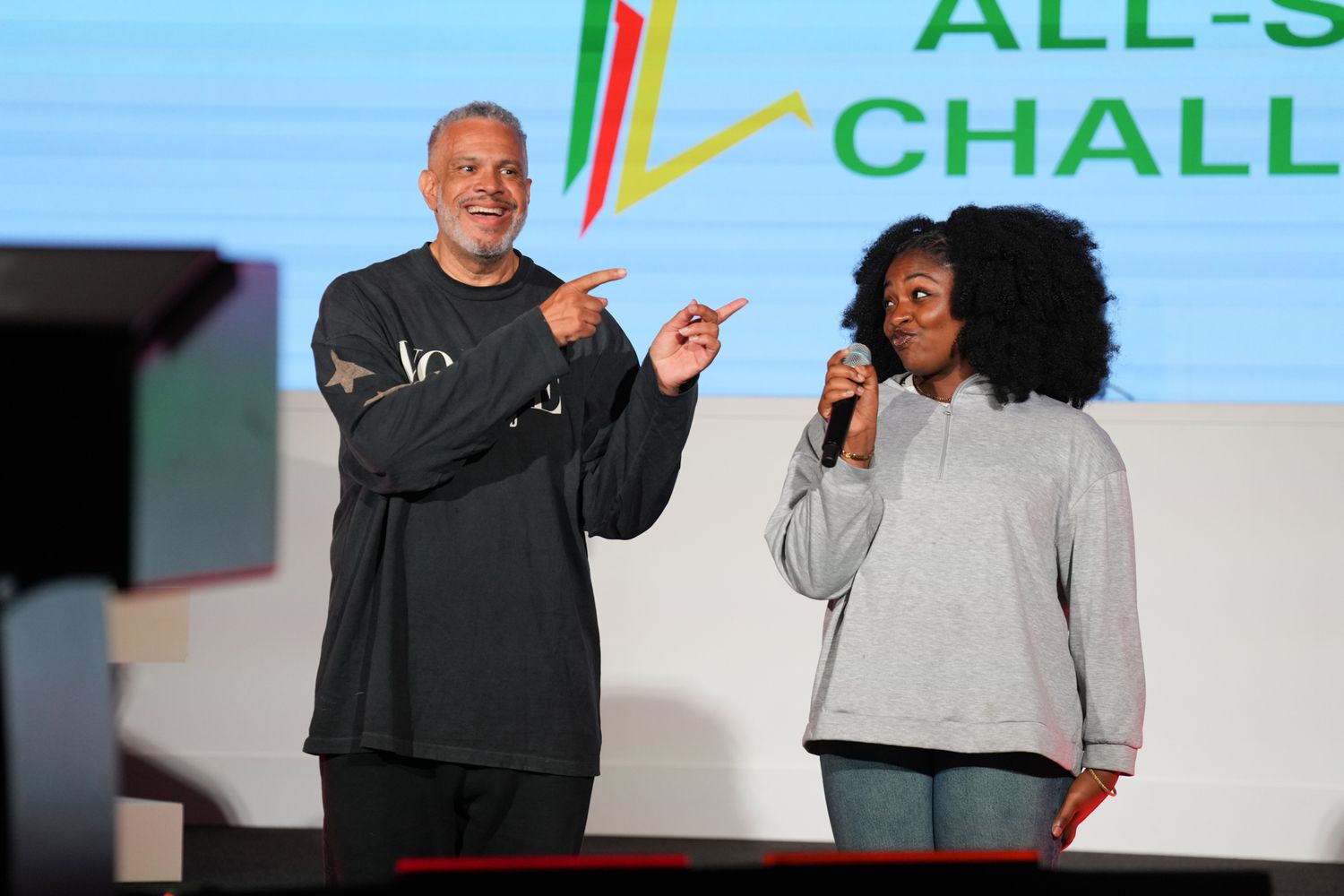 Two people on stage at an All-Star Challenge event, one pointing while the other holds a microphone.
