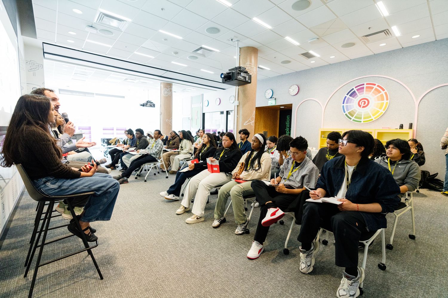 Students sitting in rows listen attentively to a speaker during an indoor classroom or seminar session.