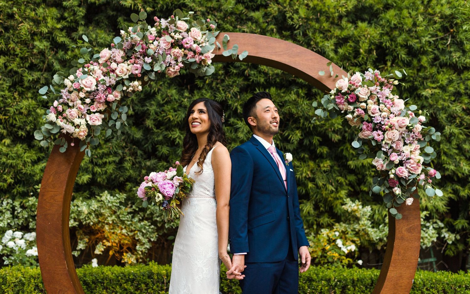 A wedding couple poses under a circular floral arch with pink and white blooms against lush greenery.