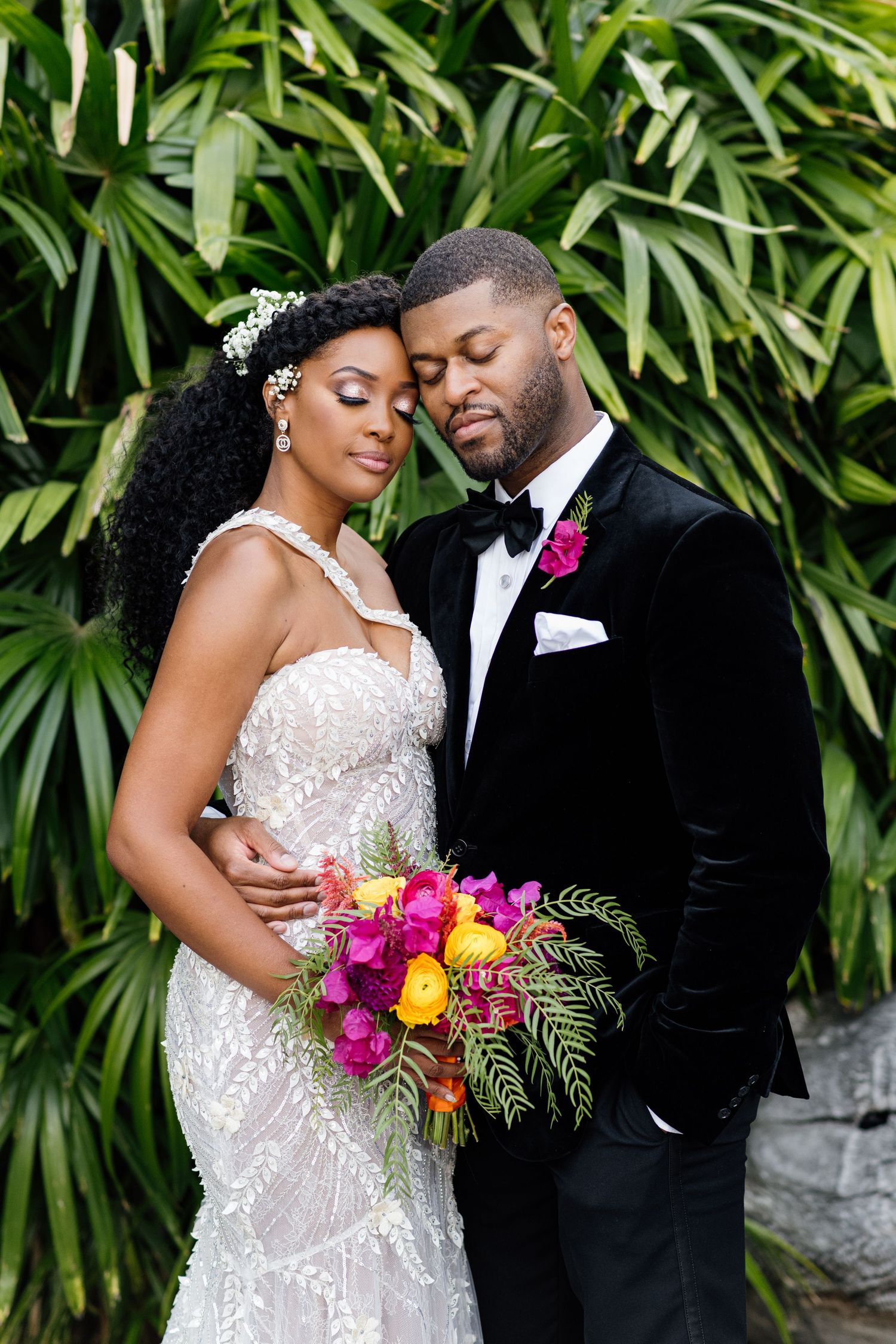 Wedding couple embraces against tropical foliage with vibrant floral bouquet.