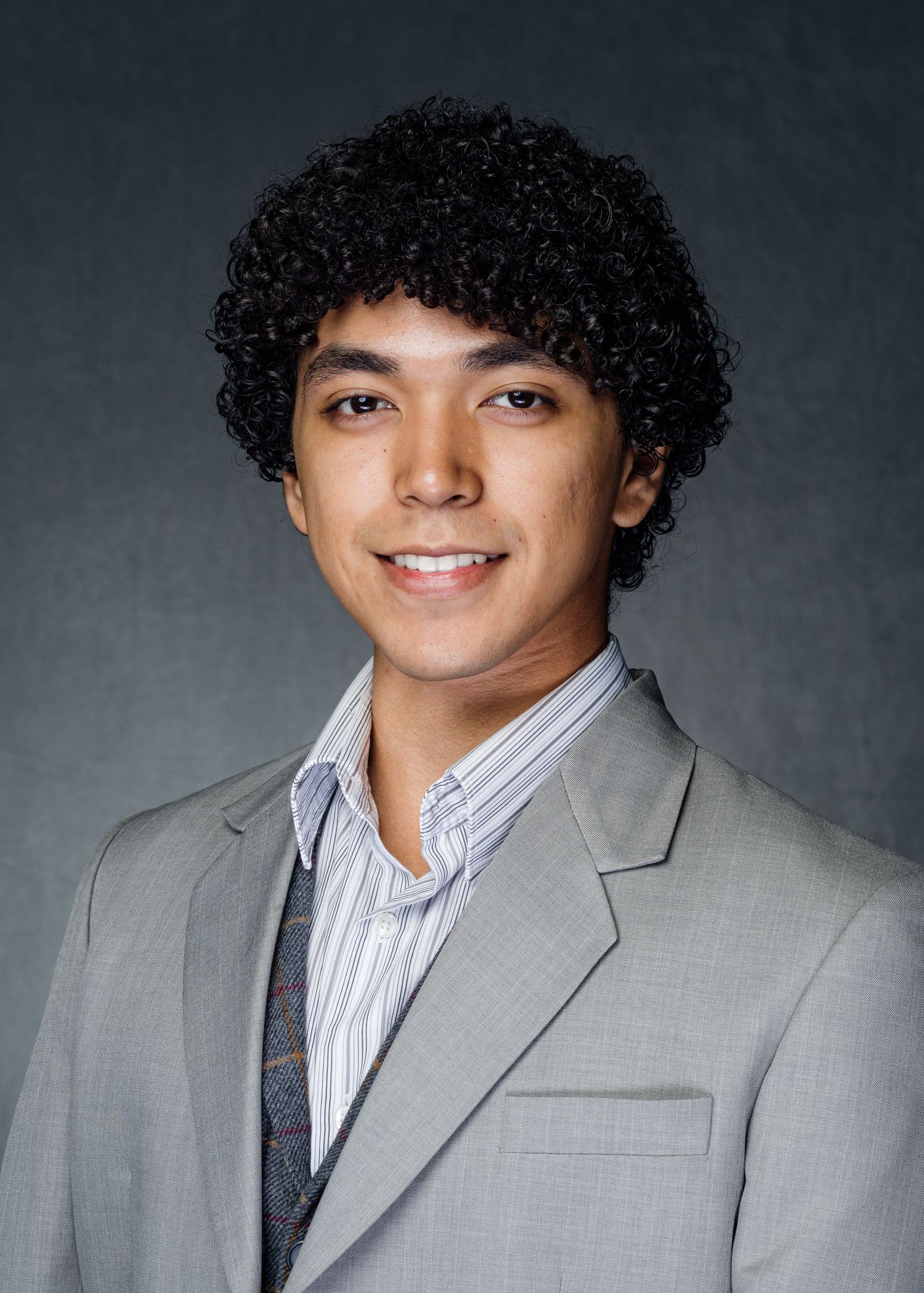 Professional headshot of someone wearing a gray suit jacket and striped shirt against a dark background.