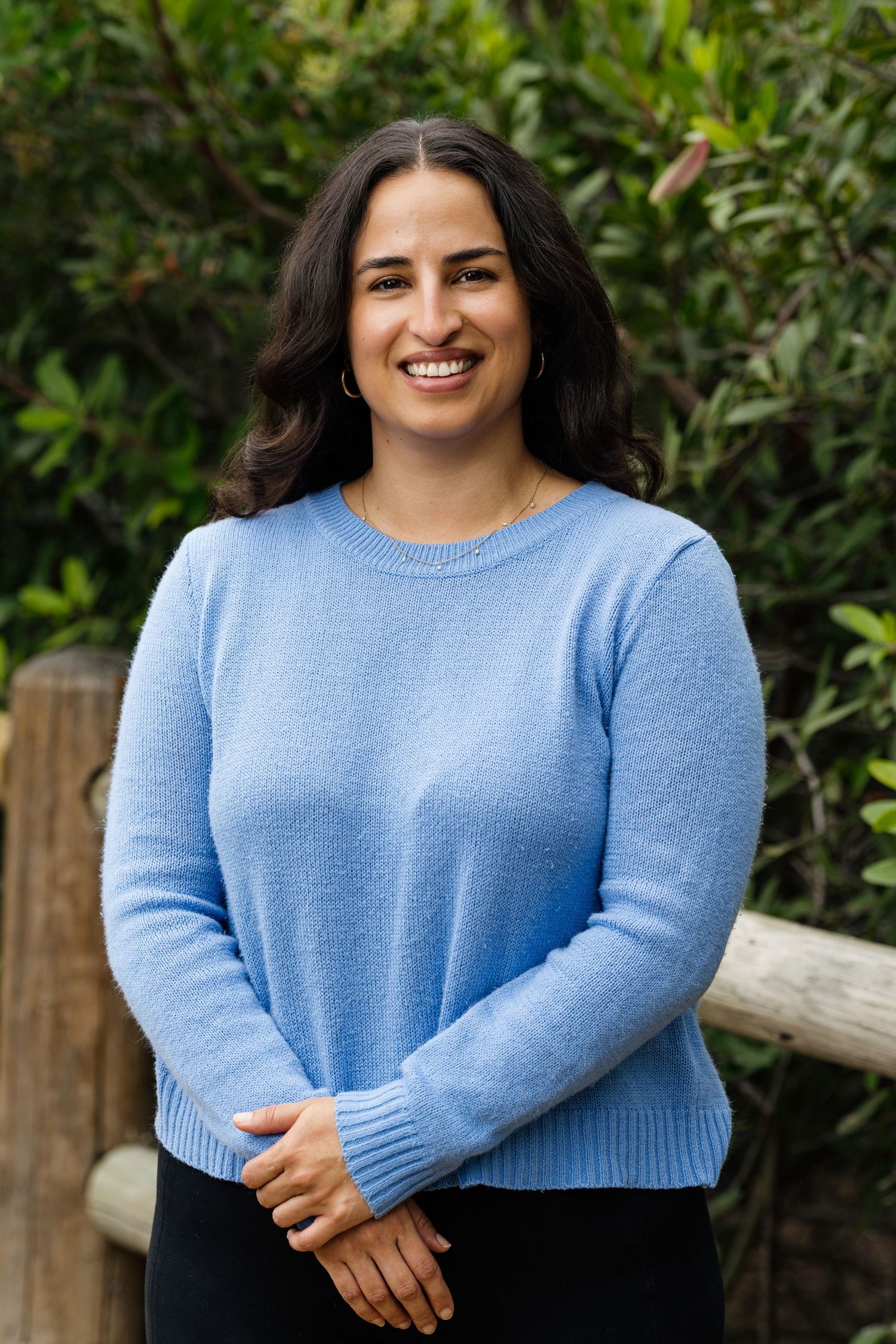 A smiling young woman in a light blue sweater poses outdoors with green foliage in the background.