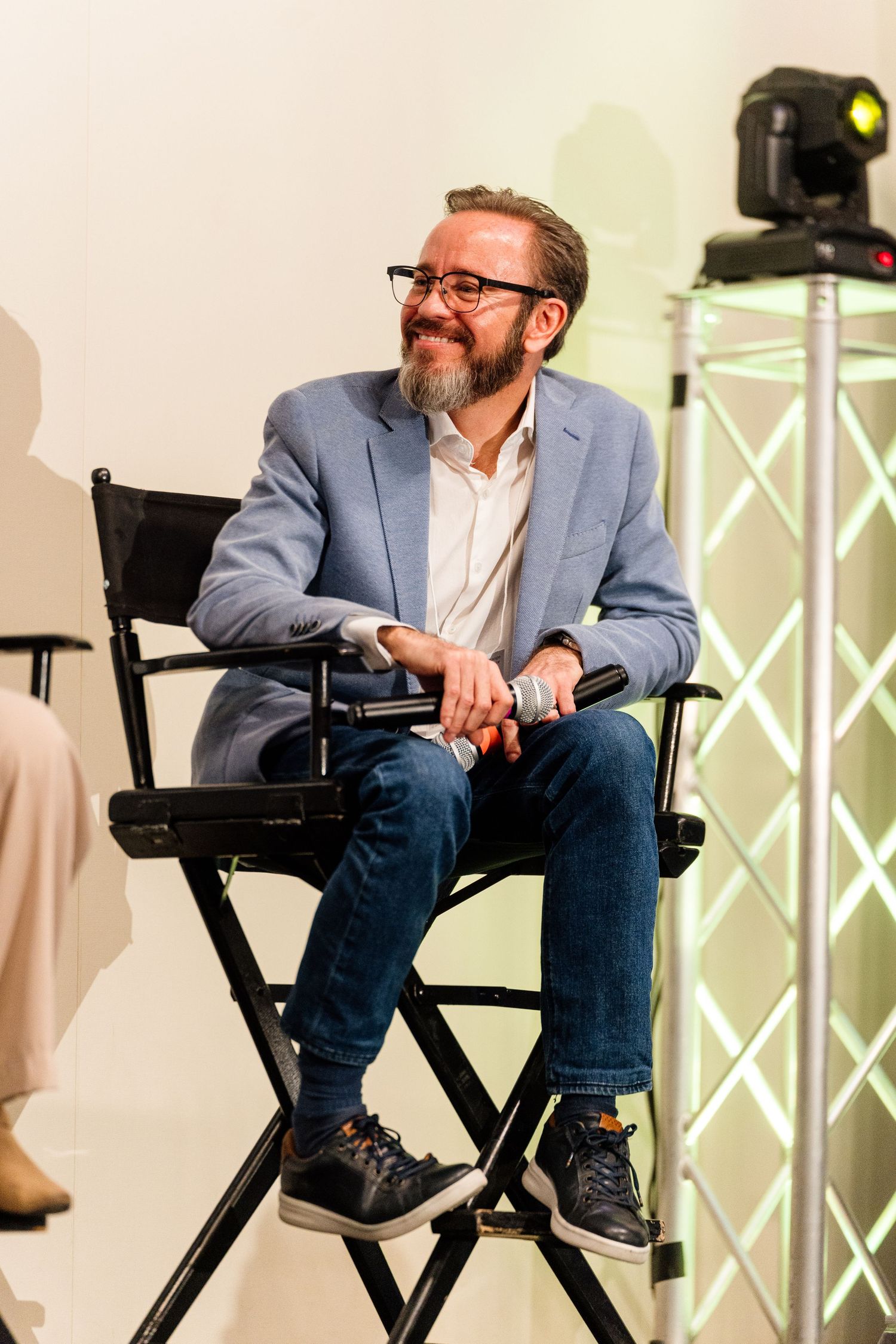 Speaker seated in director's chair wearing blue blazer during conference presentation.