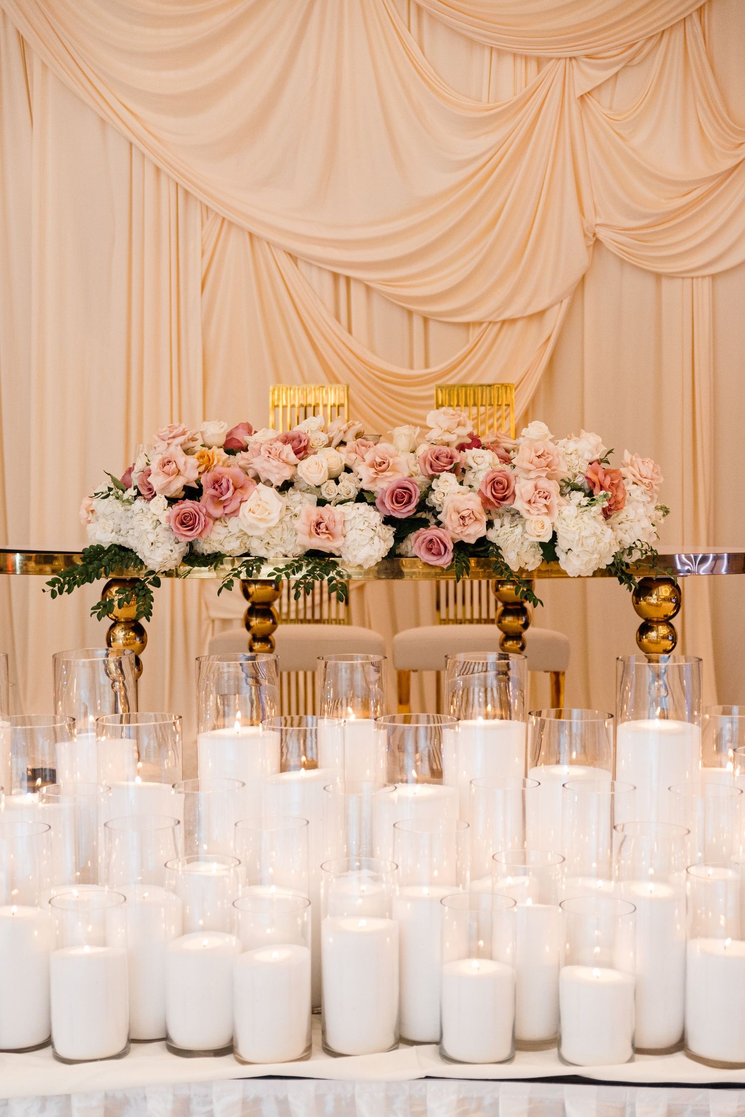 Elegant wedding reception with white pillar candles, pink and white floral centerpiece, gold chairs, and ivory draped backdrop.