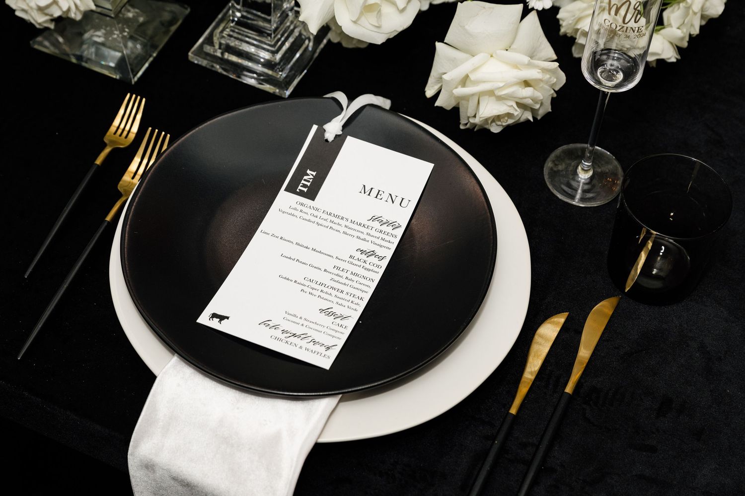 Elegant black and white place setting with gold cutlery, menu card, and white floral arrangement on dark tablecloth.