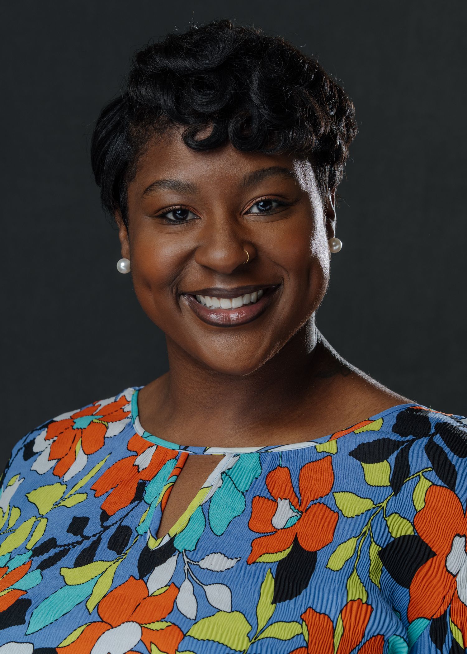 Professional headshot of a smiling woman wearing a colorful floral blouse against a dark background.