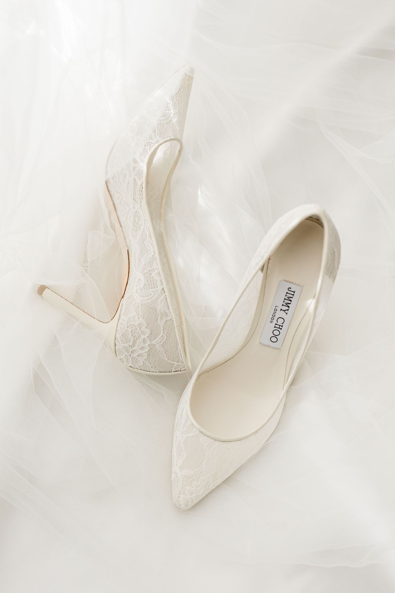 Elegant Jimmy Choo white lace pointed-toe stiletto heels on ivory tulle fabric, perfect bridal wedding shoes.