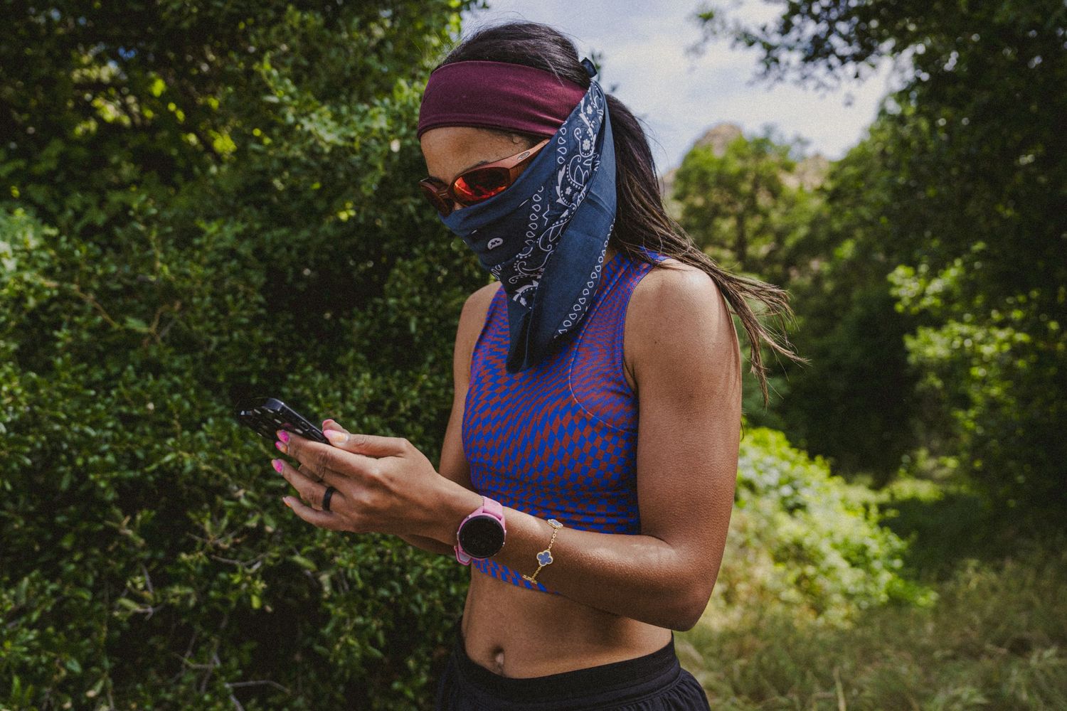 Woman in purple crop top and face mask checks her phone on a trail surrounded by green trees.