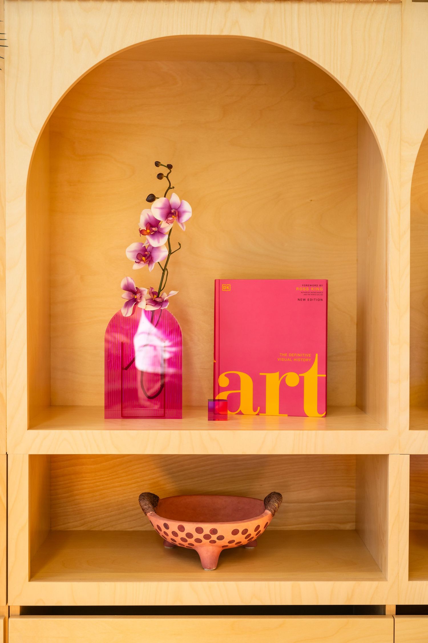 Wooden shelving unit with pink books and orchid flowers in arched compartments.