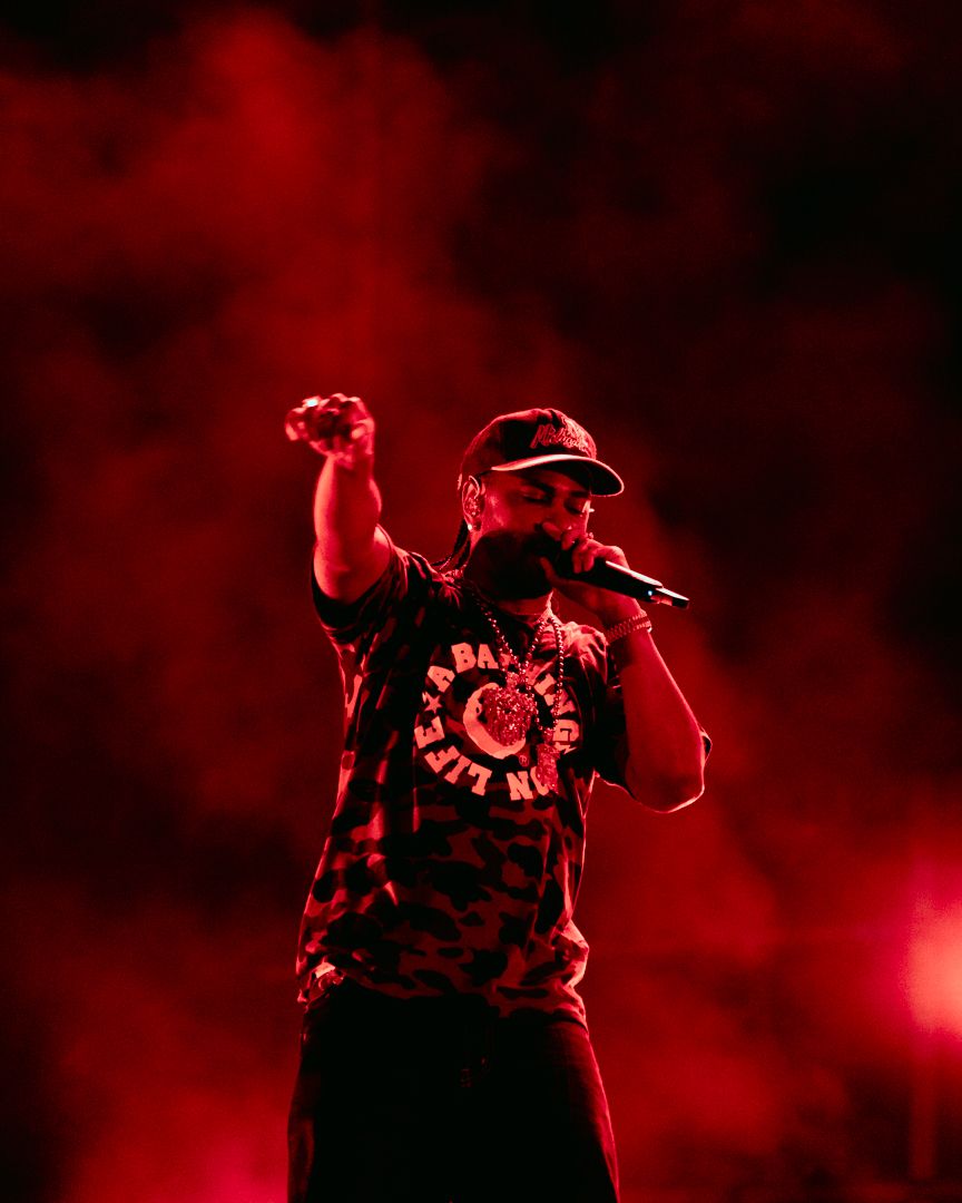 A rapper performs on stage under dramatic red lighting, raising a fist while holding a microphone in smoky atmosphere.