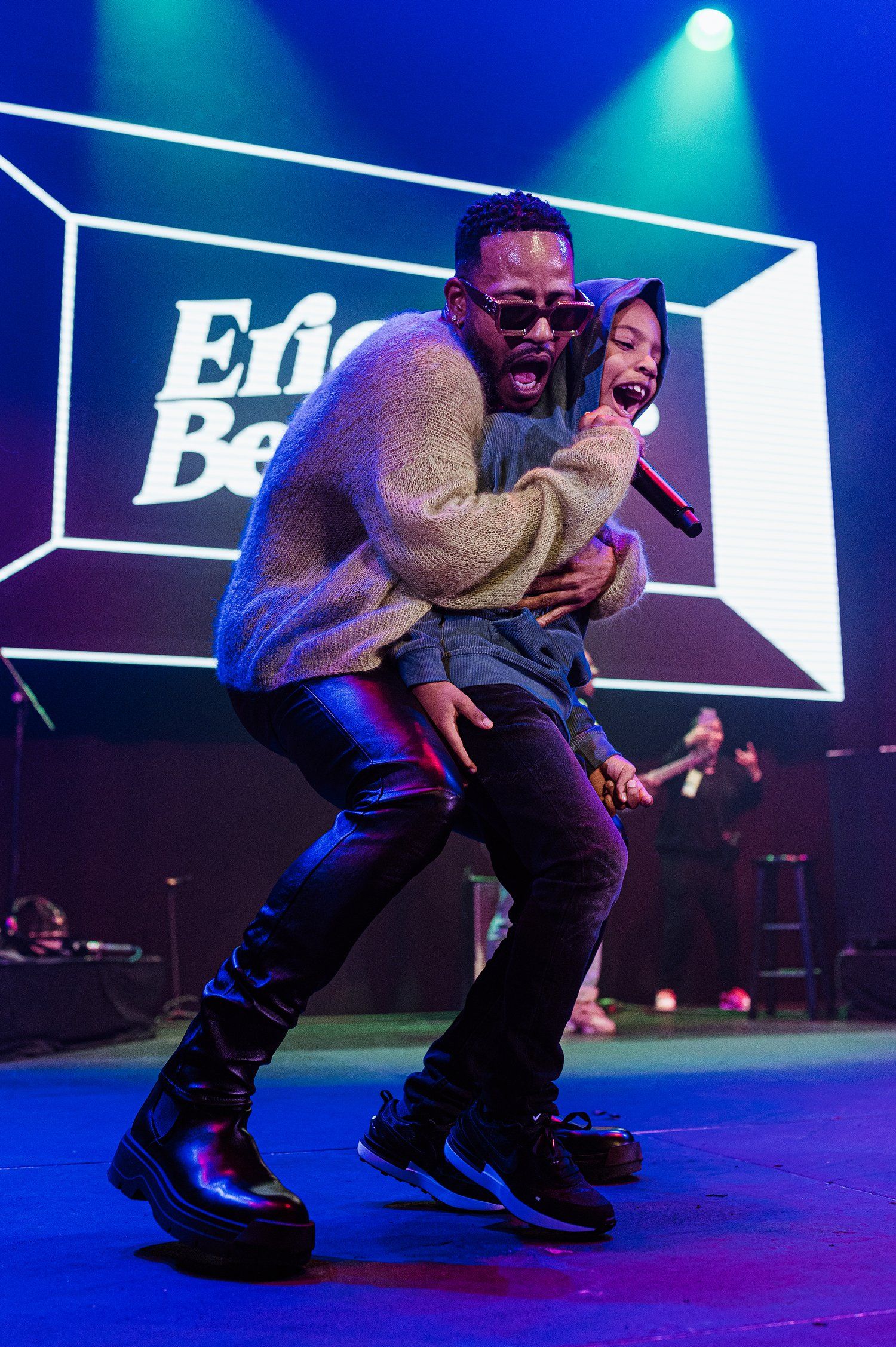 Energetic performance on blue-lit stage with geometric backdrop displaying 'Ed Be' text.