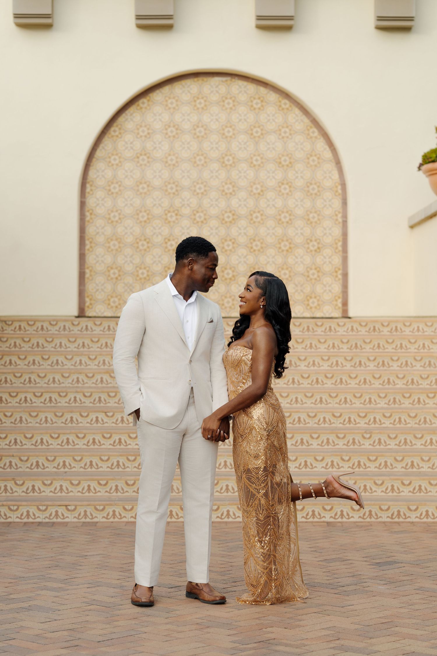 Couple in elegant white and gold attire poses against a patterned wall during a formal photoshoot.
