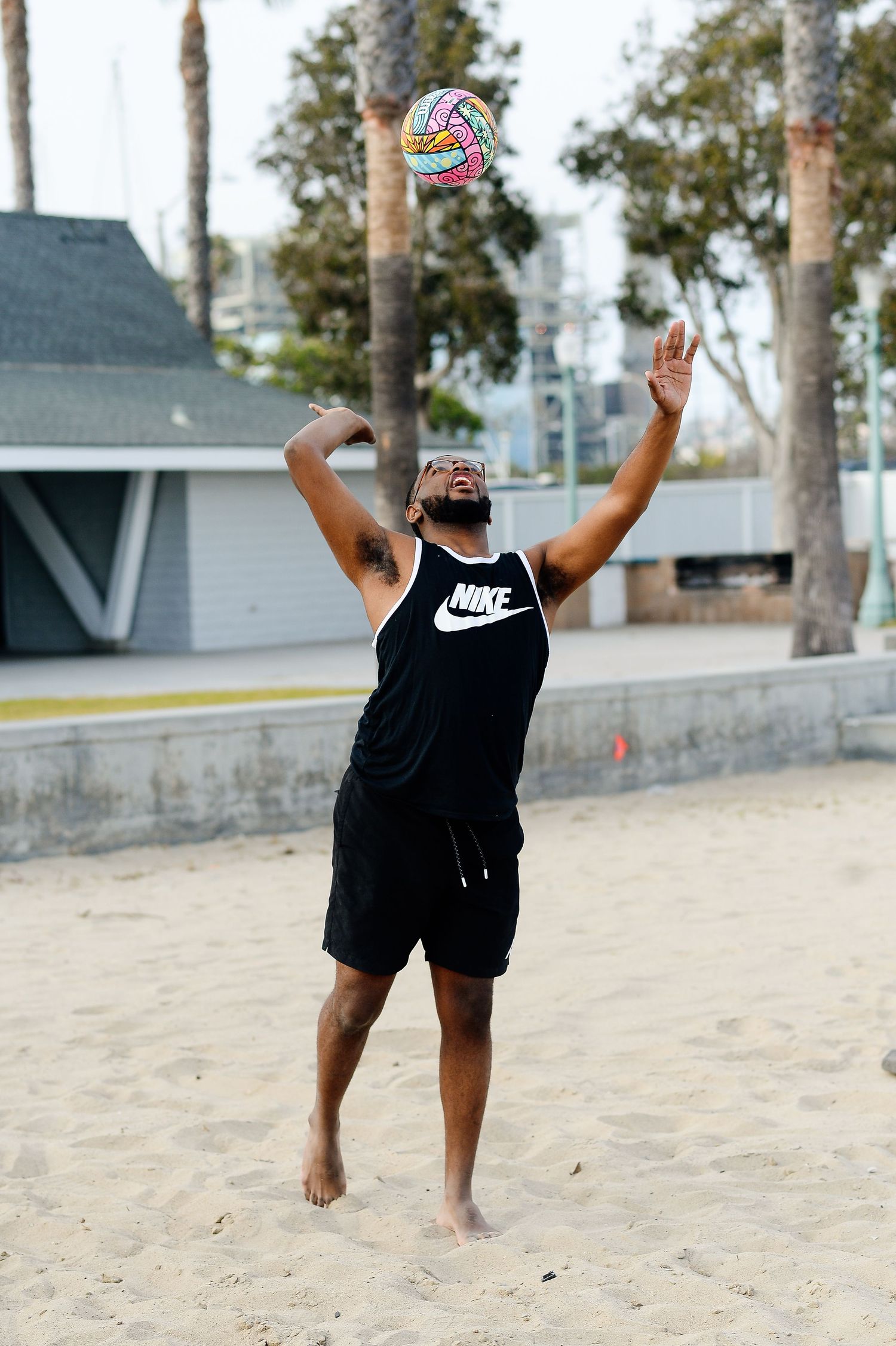Person in black Nike shirt plays volleyball on a sandy beach with palm trees in background.