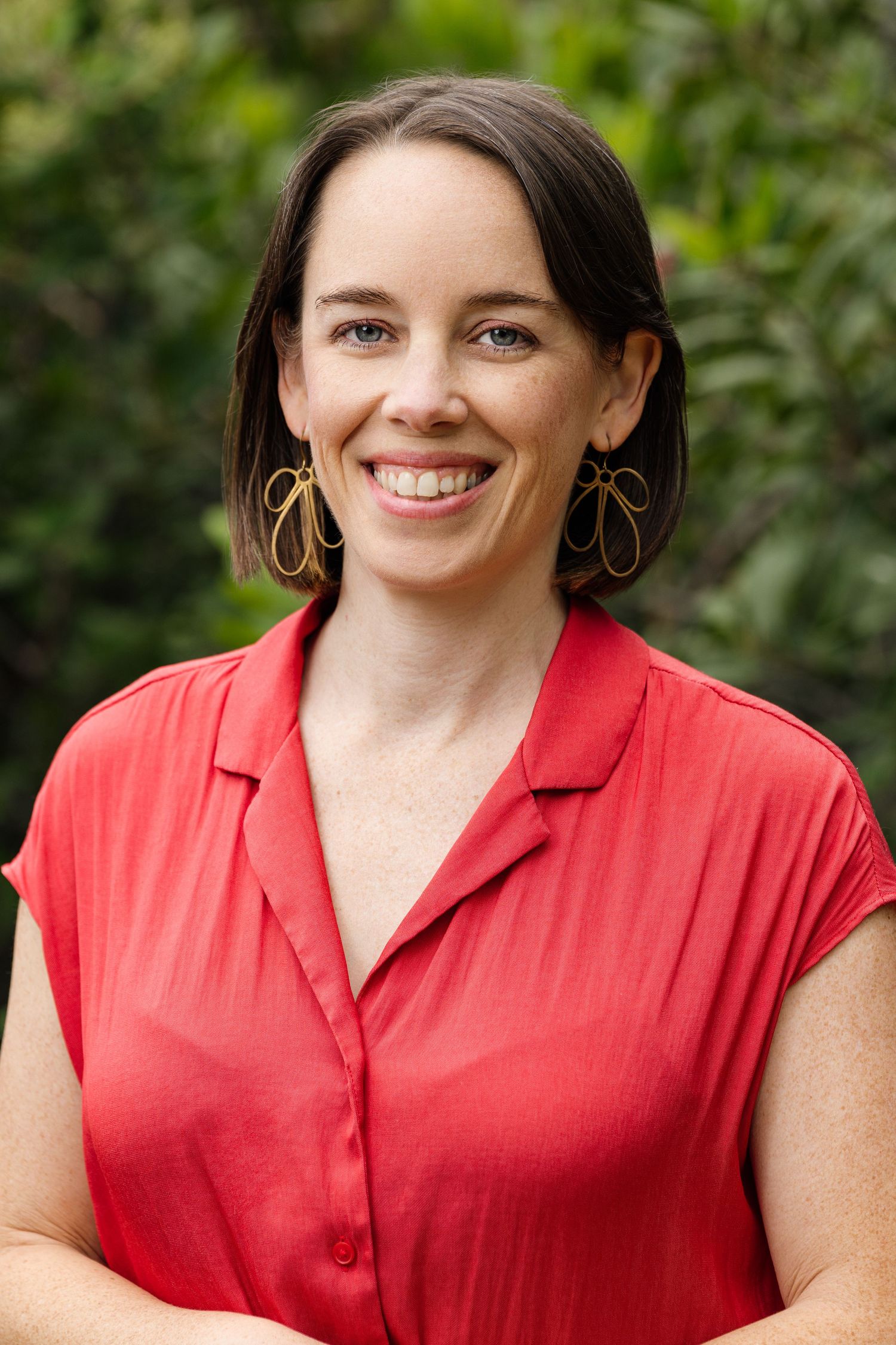 Smiling woman with dark hair wearing a red blouse and gold earrings, posing outdoors with green foliage background.
