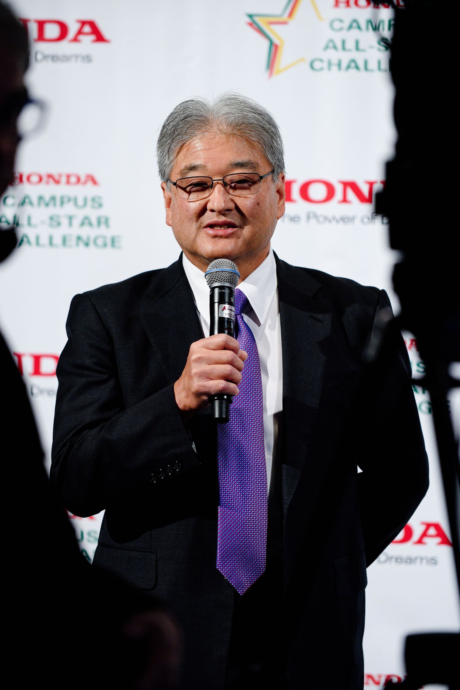 Speaker at Honda event podium wearing dark suit and purple tie.