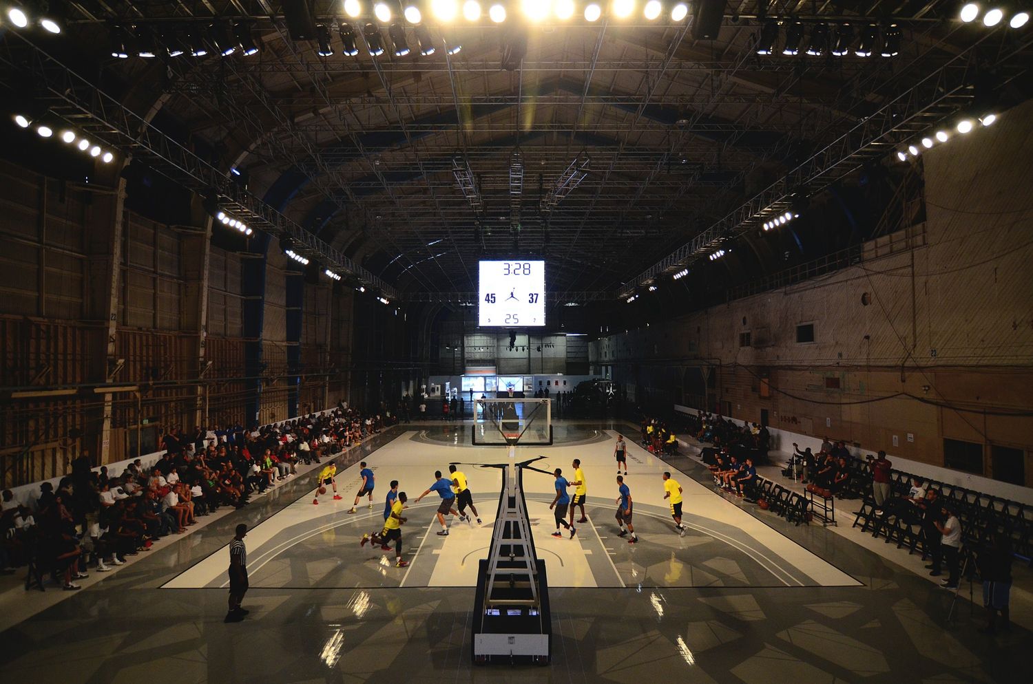 Indoor basketball court with spectators watching game under industrial lighting.