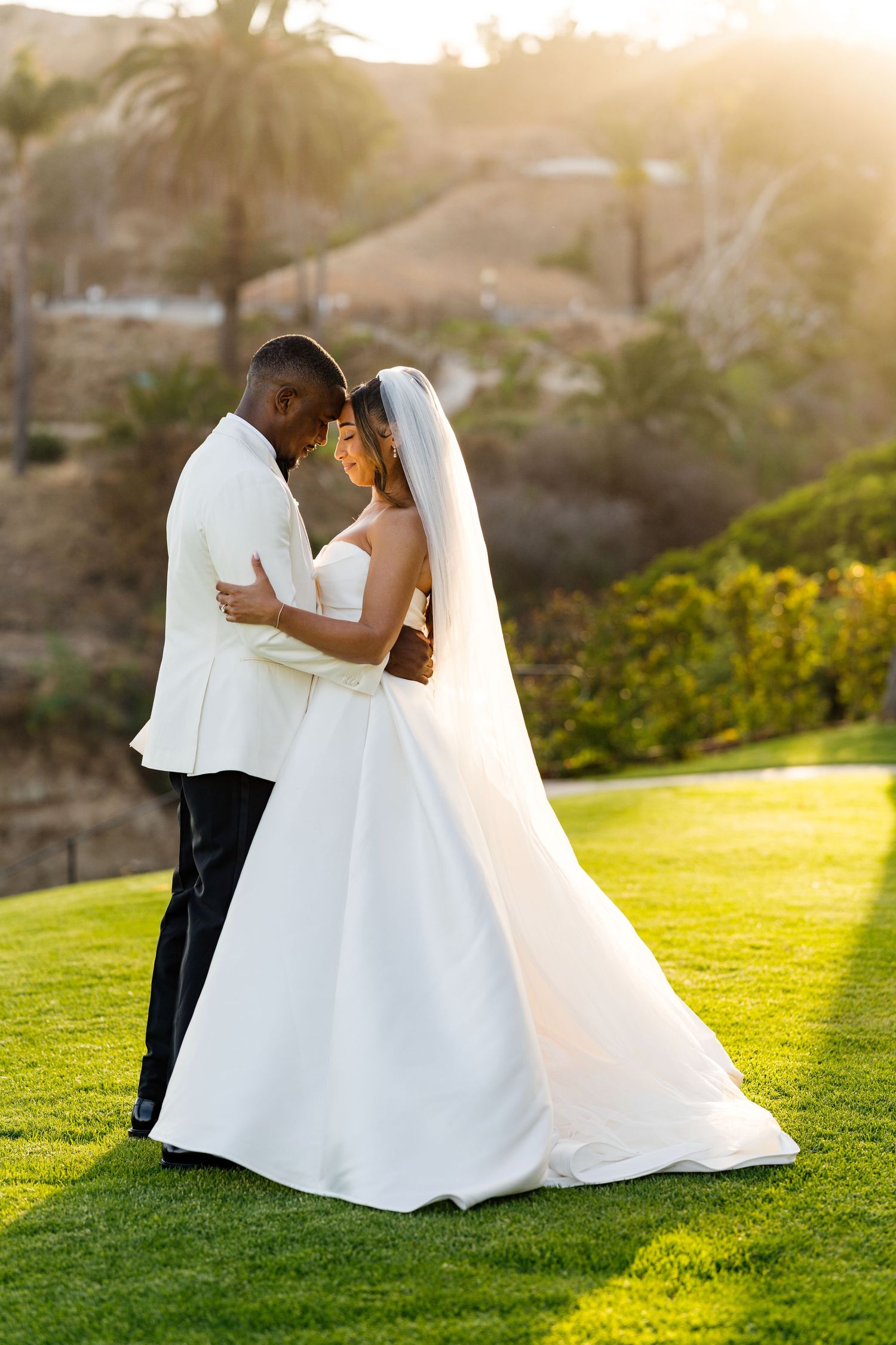 A bride and groom share a romantic embrace on a lush green lawn at golden hour during their outdoor wedding.