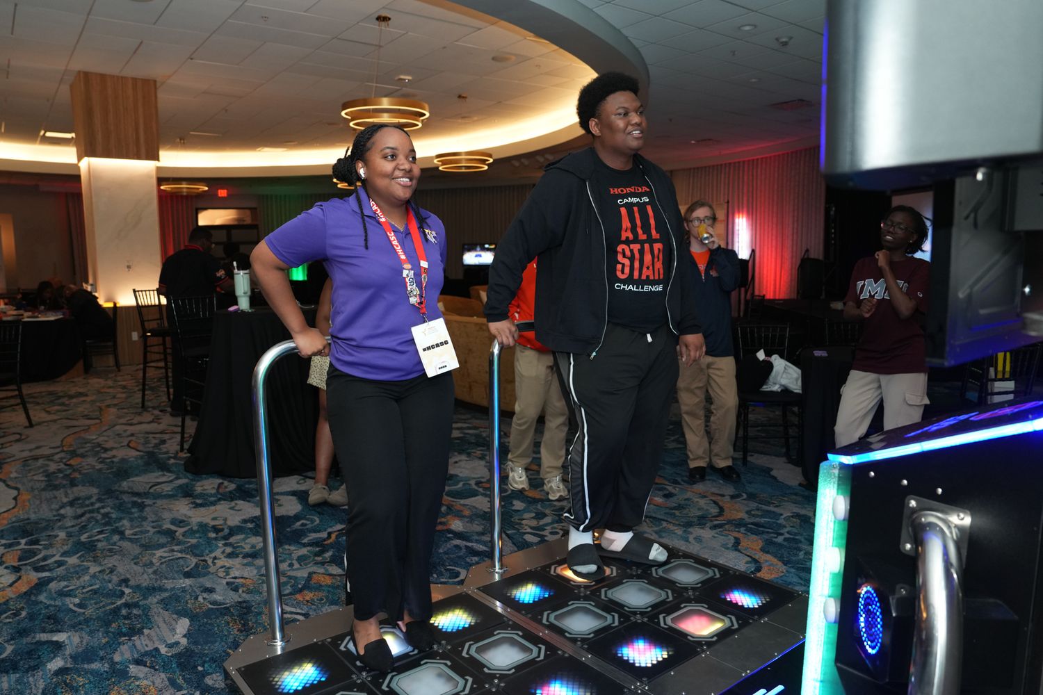 Two people playing Dance Dance Revolution at an indoor event, with colorful light-up dance pads on the floor.